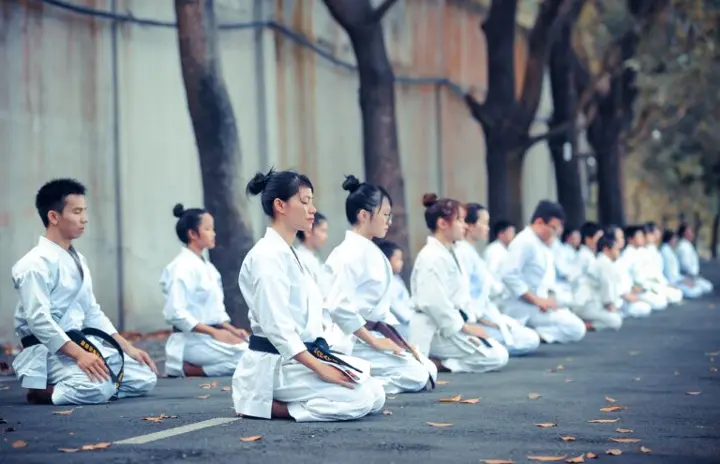 Group of martial artists in white gis meditating outdoors, focusing on discipline, mindfulness, and inner peace as part of their training.