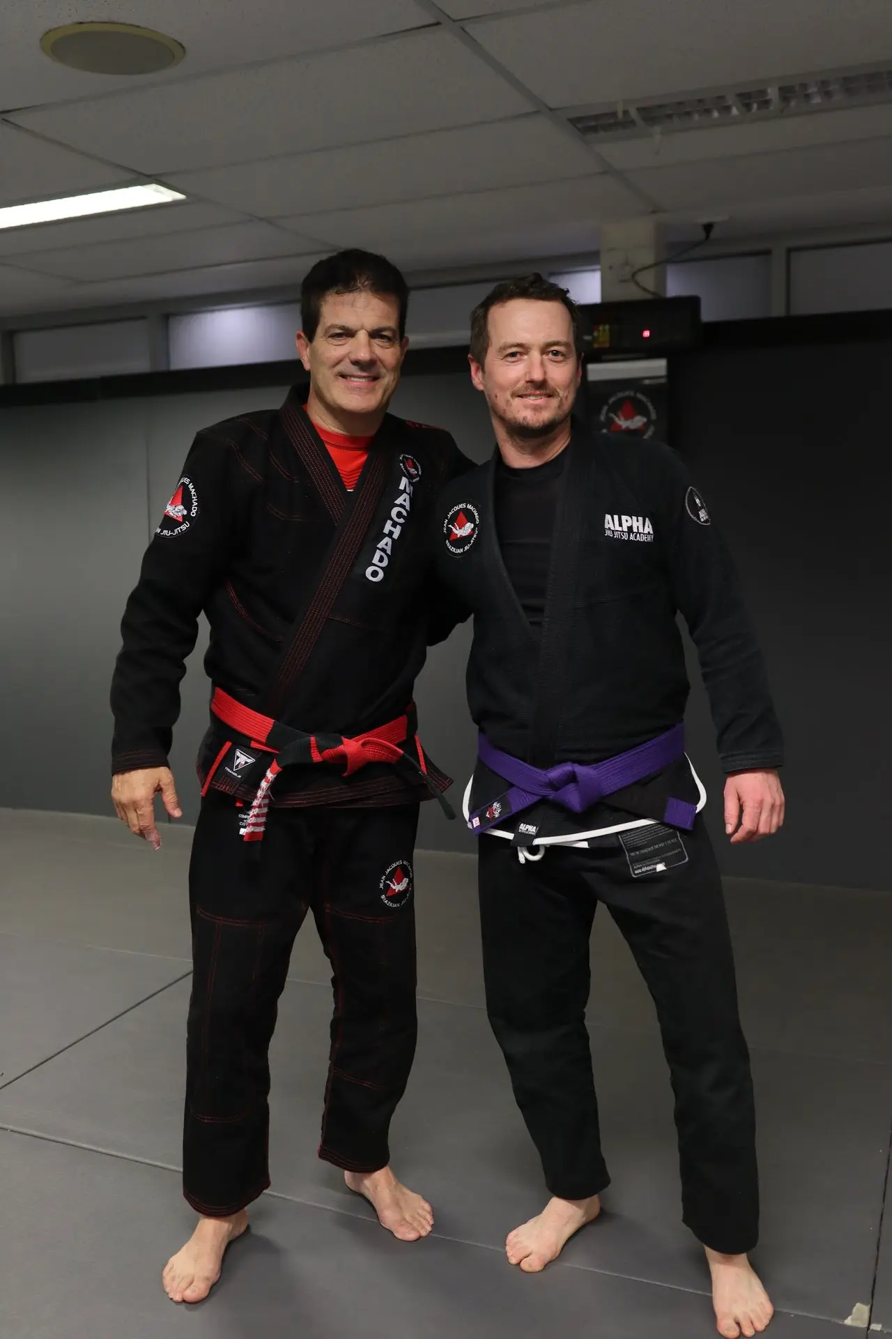 Jean-Jacques Machado posing with a purple belt student from Alpha Jiu Jitsu Academy, both in Brazilian Jiu-Jitsu gis, during a training session at the academy.