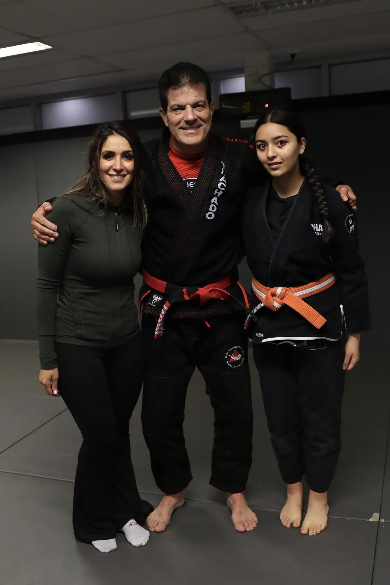 A group photo of Marie Sales, Program Director, standing next to Jean-Jacques Machado in a black gi with a red belt, and an orange belt student in a black gi at Alpha Jiu-Jitsu Academy. They are posing together, showcasing the unity and dedication in Brazilian Jiu-Jitsu training and mentorship.