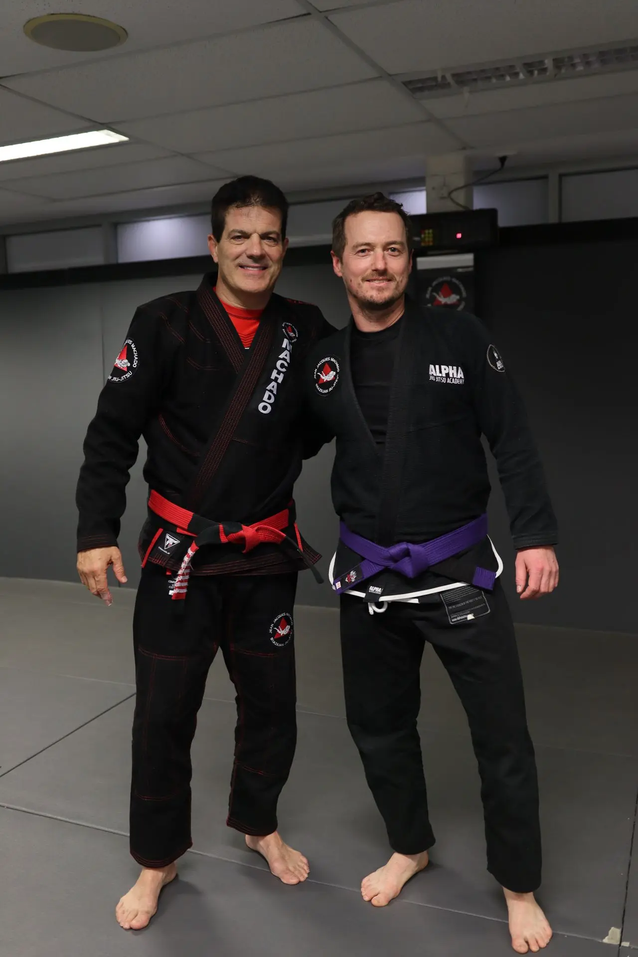 Jean-Jacques Machado, wearing a black gi and red belt, stands with a purple belt student in a black gi at Alpha Jiu-Jitsu Academy. Both individuals are smiling and standing together on the mat, symbolizing the camaraderie and progress achieved in Brazilian Jiu-Jitsu training.