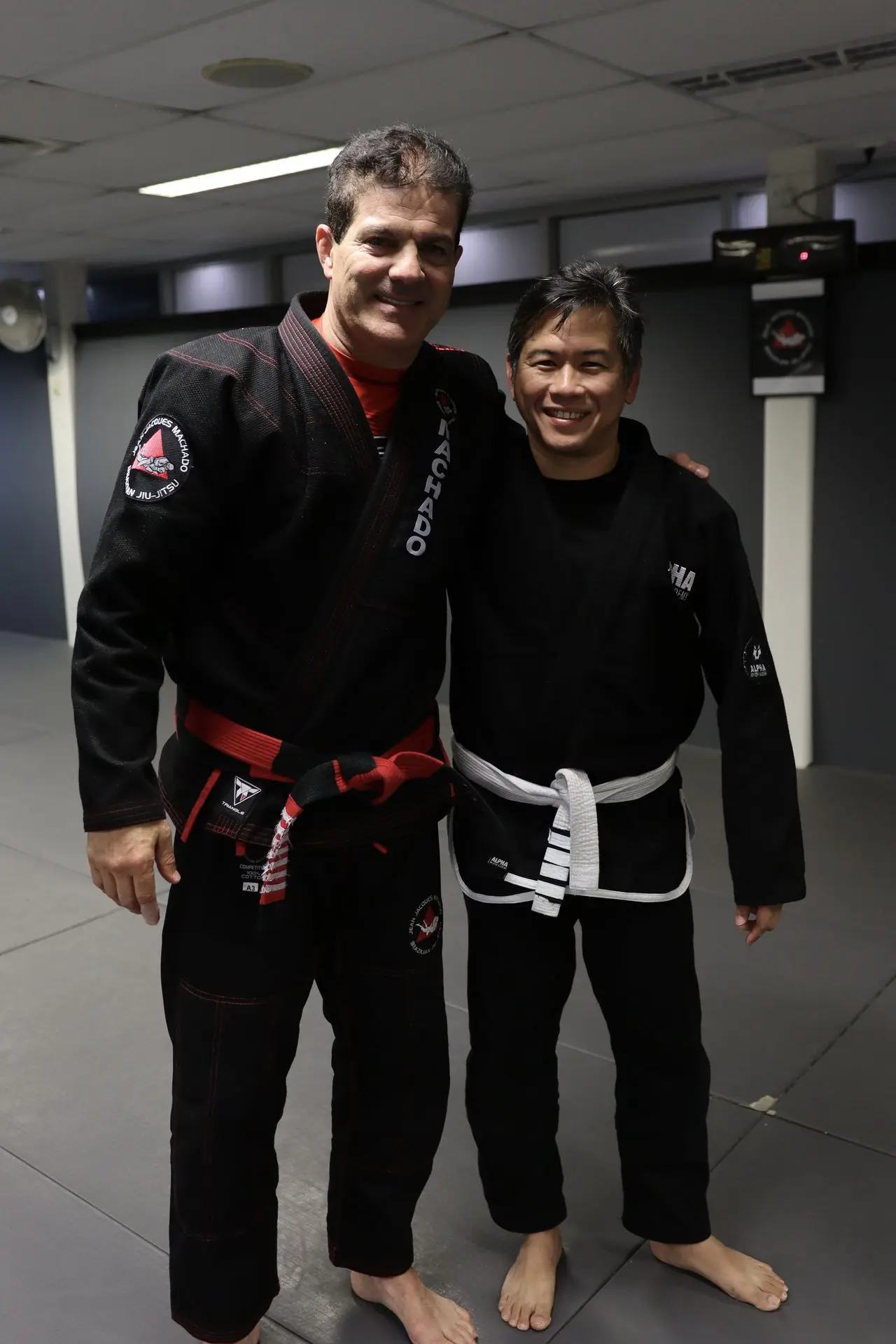 Jean-Jacques Machado, dressed in a black gi with a red belt, stands smiling next to a white belt practitioner wearing a black gi with an Alpha Jiu-Jitsu Academy patch. The image captures a moment of camaraderie and mentorship on the mats at Alpha Jiu-Jitsu Academy.