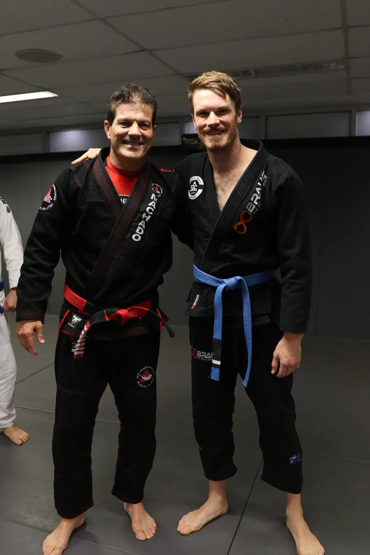 Jean-Jacques Machado, wearing a black gi and red belt, stands next to a blue belt student at Alpha Jiu-Jitsu Academy. Both are smiling and standing arm-in-arm, showcasing camaraderie and mentorship in Brazilian Jiu-Jitsu training. The student wears a black gi with a blue belt, representing their level in BJJ. The background shows a typical training facility setting, emphasizing the supportive and skill-driven environment of the academy.