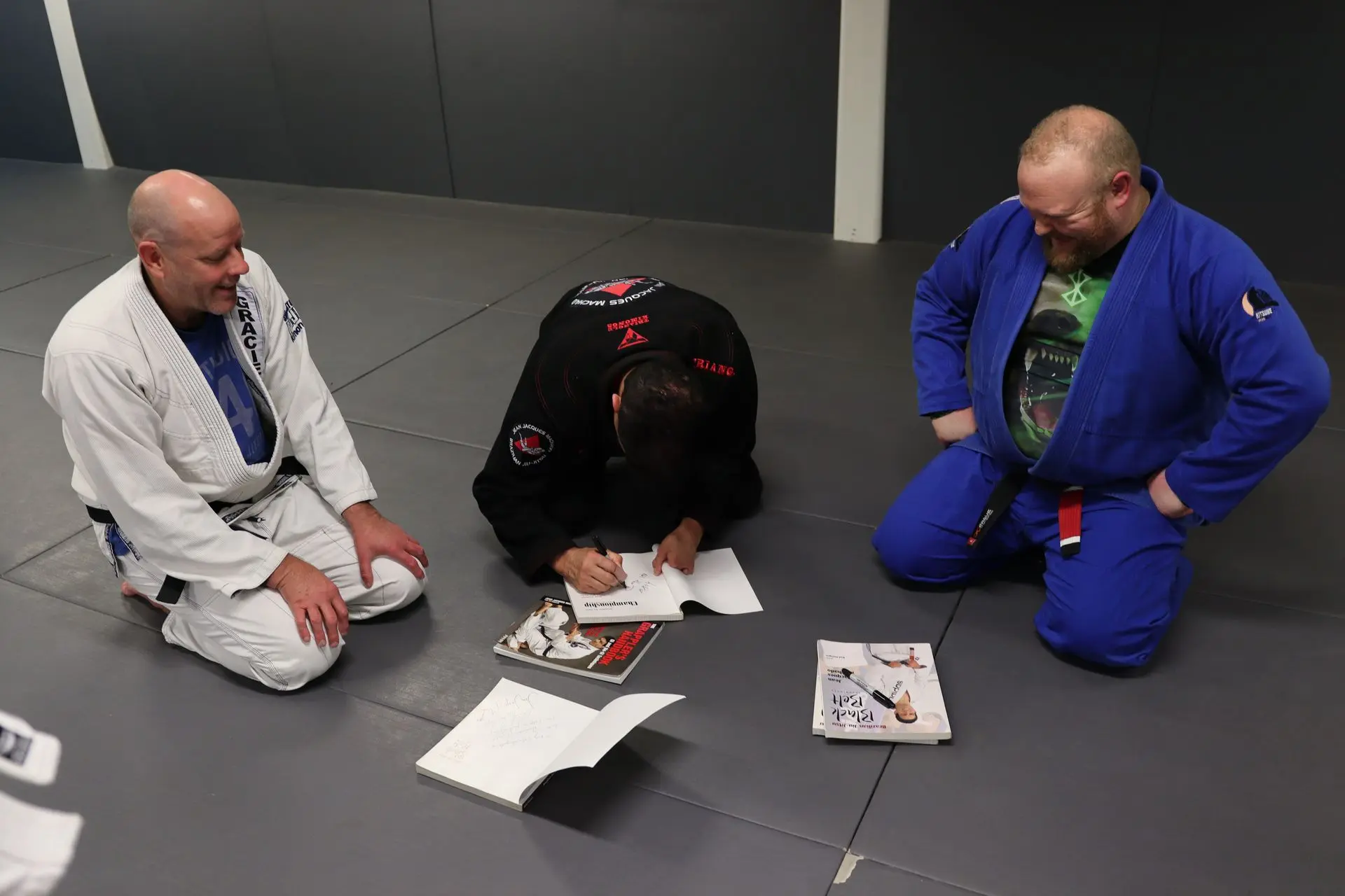 Jean-Jacques Machado, wearing a black gi, is seated and signing books for students at Alpha Jiu-Jitsu Academy. Two students, one wearing a white gi and the other in a blue gi, kneel beside him, observing with smiles and interest. Open books and a closed book featuring martial arts content are visible on the mat, highlighting the knowledge-sharing and mentorship atmosphere in Brazilian Jiu-Jitsu training. The setting captures the engaging and respectful student-teacher interaction within the academy's training facility.