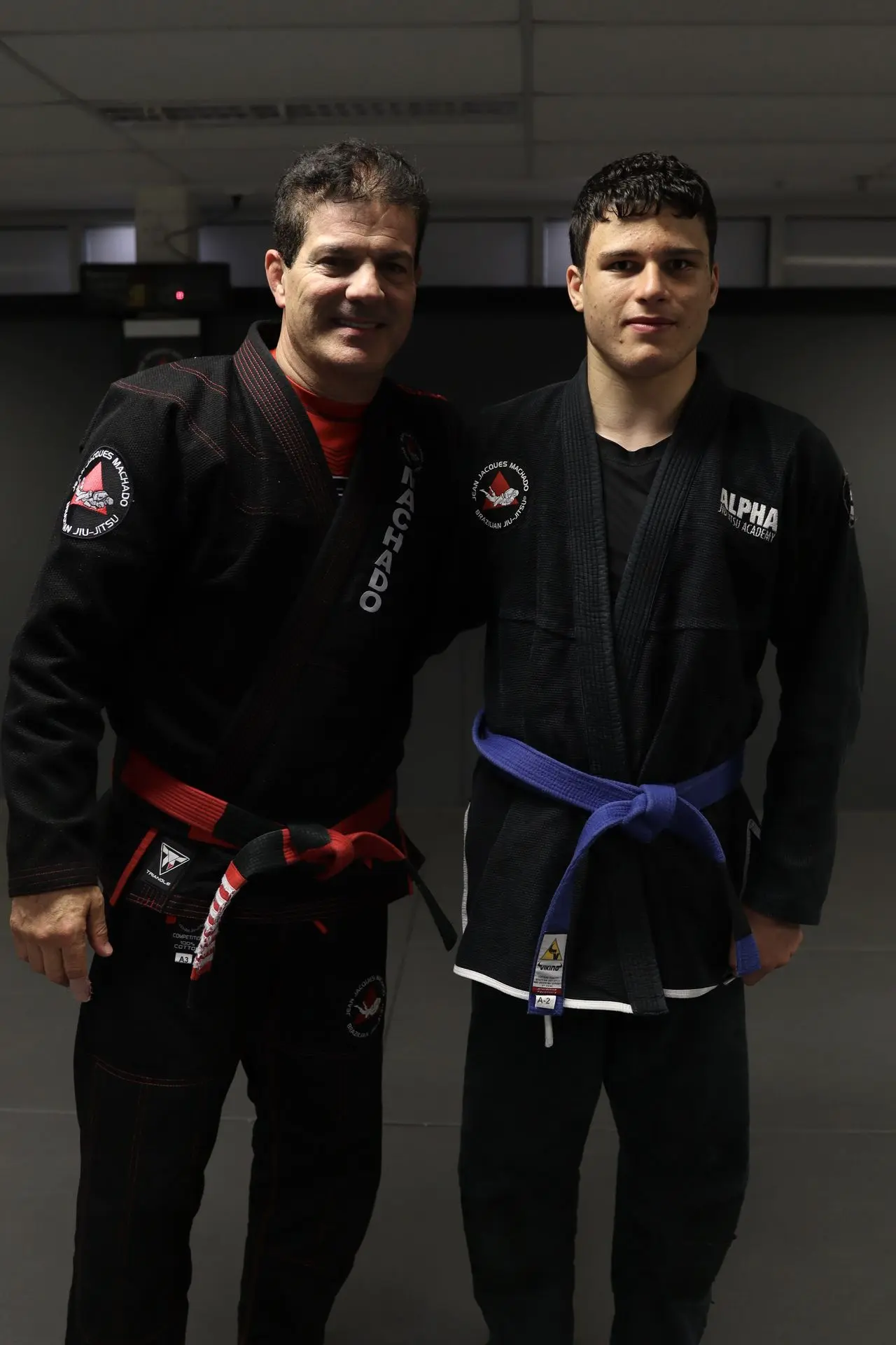 Jean-Jacques Machado, wearing a black gi with red belt, stands next to a blue belt practitioner dressed in a black gi with an Alpha Jiu-Jitsu Academy patch, posing for a photo at Alpha Jiu-Jitsu Academy. The scene emphasizes mentorship and progression in Brazilian Jiu-Jitsu training.