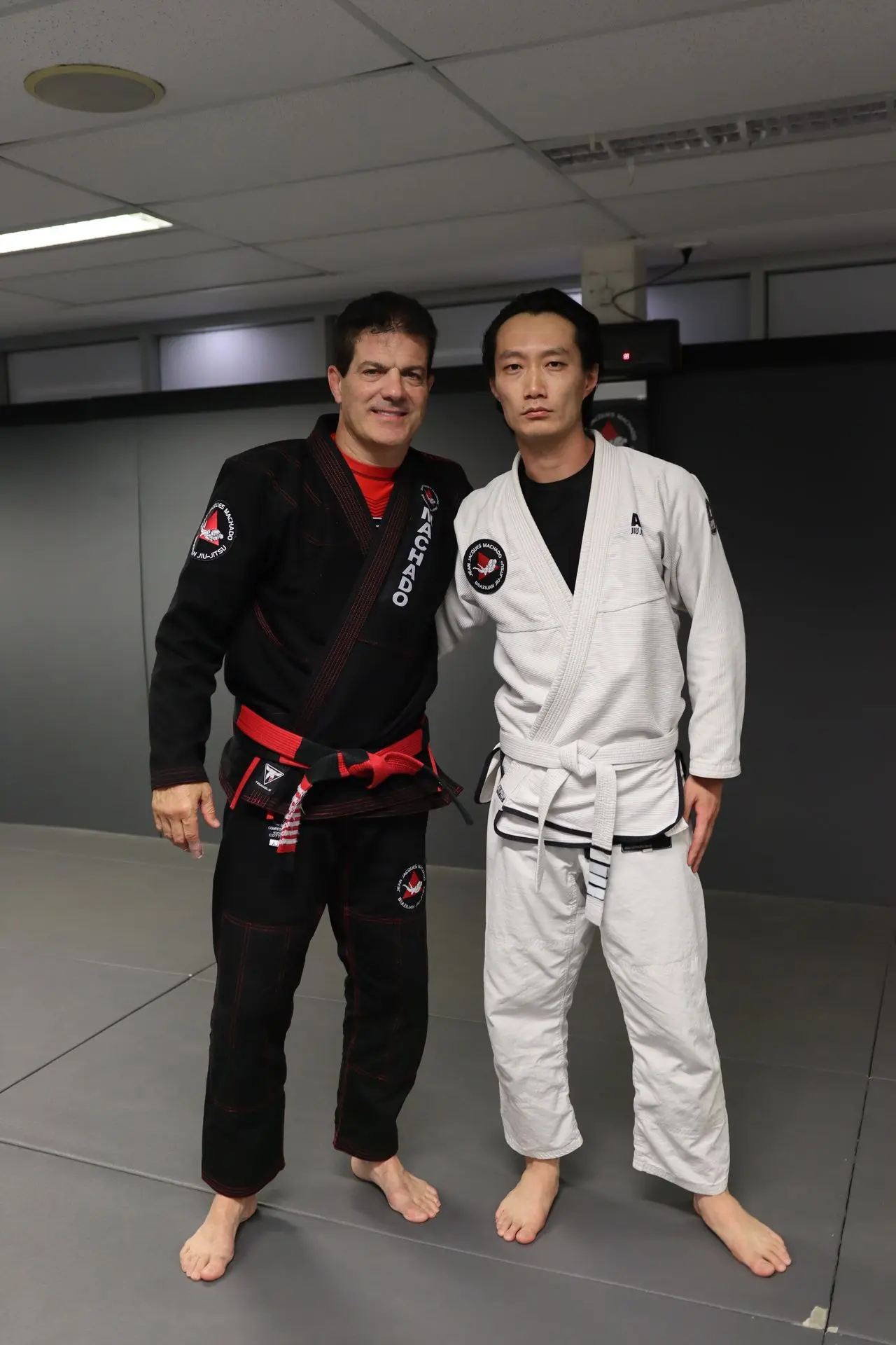 Jean-Jacques Machado, in a black gi with a red belt, stands next to a BJJ student wearing a white gi with a white belt at Alpha Jiu Jitsu Academy, posing for a photo on the academy mats.