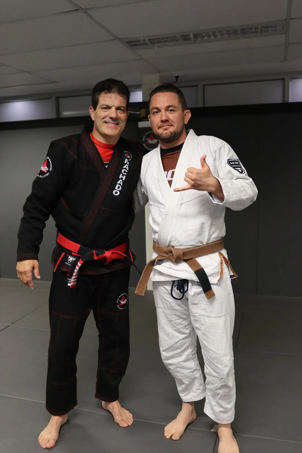 Jean-Jacques Machado stands beside a brown belt student at Alpha Jiu-Jitsu Academy. The student, dressed in a white gi, smiles and gives a shaka hand gesture, symbolizing the positive atmosphere of the academy. Both individuals stand on the mat, representing their dedication to Brazilian Jiu-Jitsu training and community spirit.