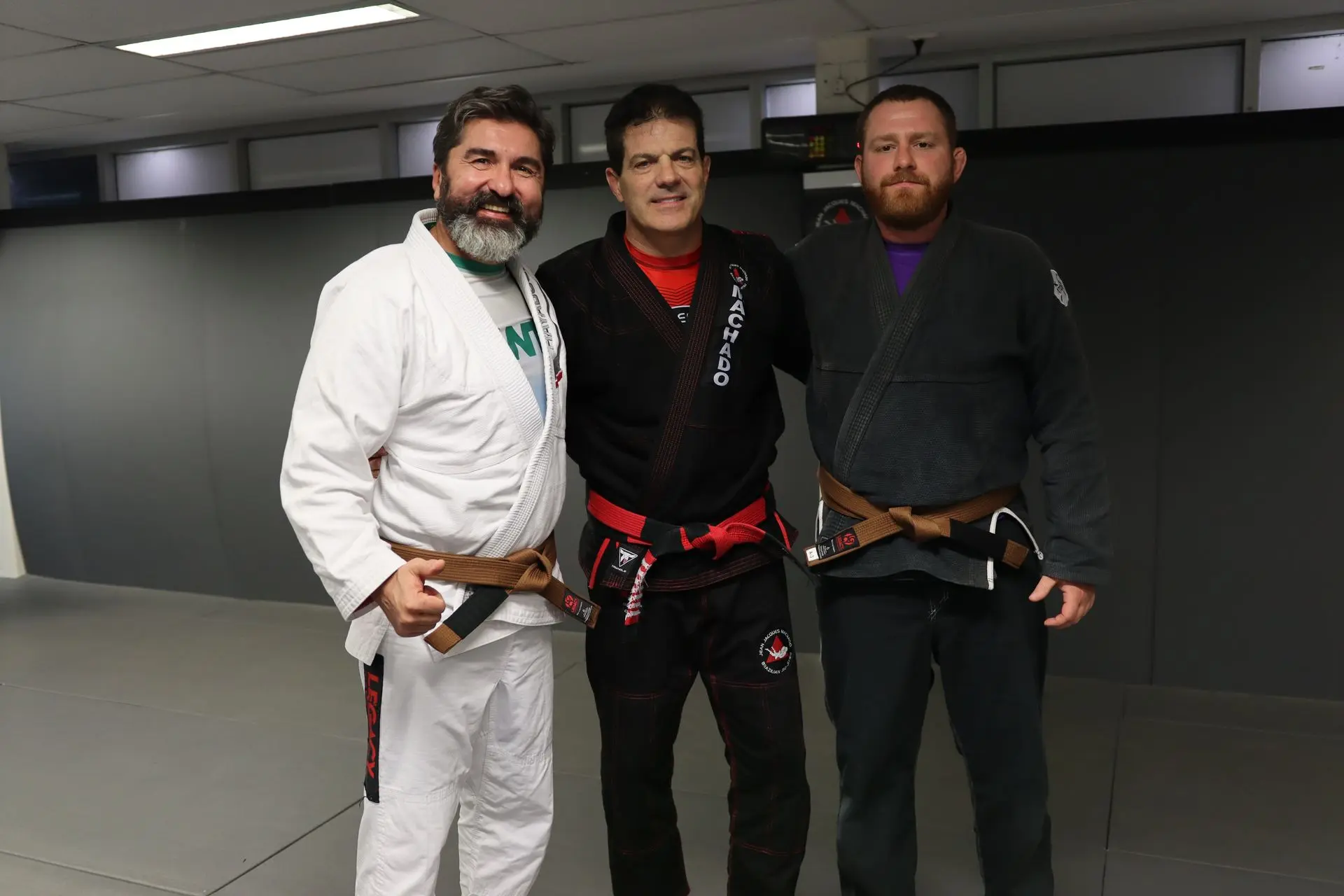 Jean-Jacques Machado stands between two brown belt students at Alpha Jiu-Jitsu Academy. The student on the left wears a white gi and the student on the right wears a black gi. All three individuals smile and pose together on the mat, showcasing their dedication and camaraderie in Brazilian Jiu-Jitsu training.