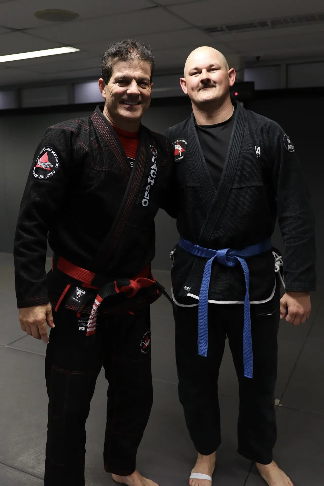 Jean-Jacques Machado, wearing a black gi and red belt, poses next to a BJJ practitioner in a black gi with a blue belt at Alpha Jiu Jitsu Academy. Both are standing on the training mats during a martial arts gathering.