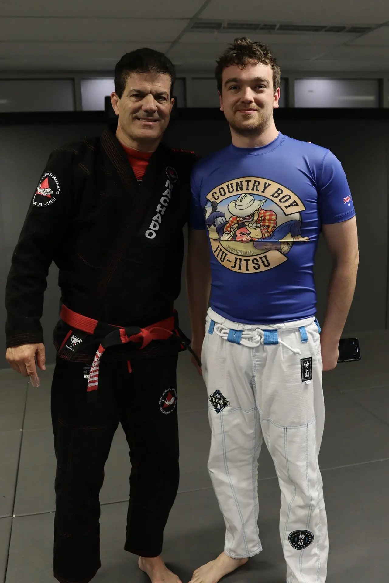 Jean-Jacques Machado standing with a BJJ student wearing a blue rash guard with a "Country Boy Jiu-Jitsu" design, both posing at a Brazilian Jiu-Jitsu academy training facility.