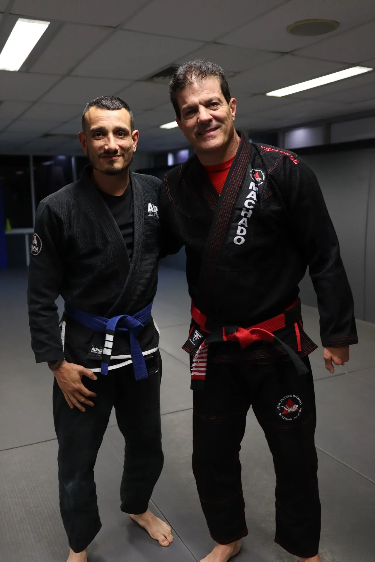 Jean-Jacques Machado, wearing a black gi with a red belt, posing next to a BJJ student in a black gi with a blue belt. The two stand together at Alpha Jiu Jitsu Academy.