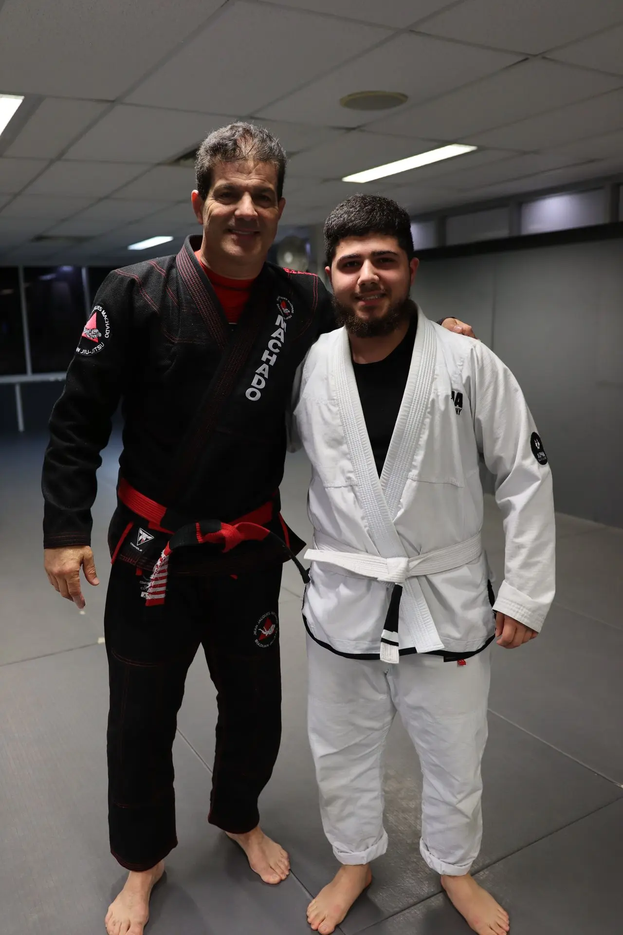 Jean-Jacques Machado, wearing a black gi and red belt, stands with a BJJ student dressed in a white gi and white belt at Alpha Jiu Jitsu Academy, posing together on the training mats.