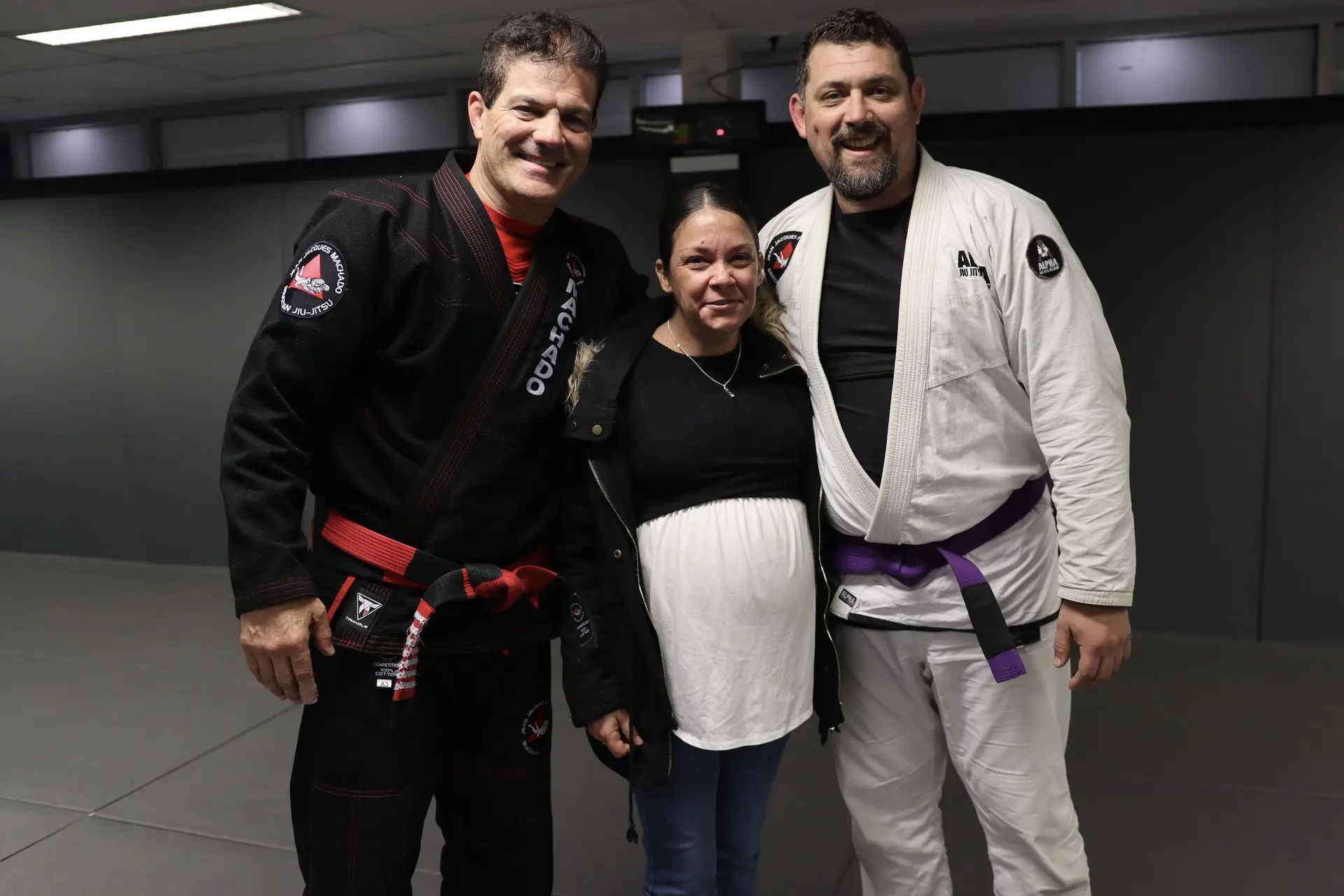 Jean-Jacques Machado, wearing a black gi with a red belt, stands with a purple belt jiu-jitsu practitioner in a white gi and a guest in a white top and black jacket at Alpha Jiu-Jitsu Academy. The group is smiling and embracing, showcasing the community and welcoming spirit within the academy.