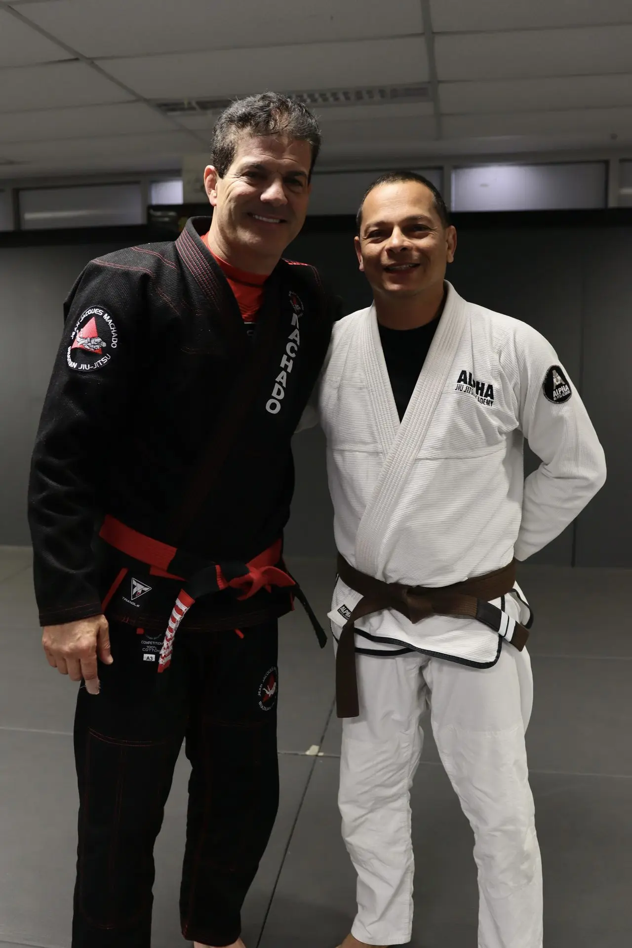 Jean-Jacques Machado, wearing a black gi and red belt, stands beside a brown belt BJJ practitioner in a white gi at Alpha Jiu Jitsu Academy. They are smiling and posing on the training mats, capturing a moment during a martial arts event or gathering.