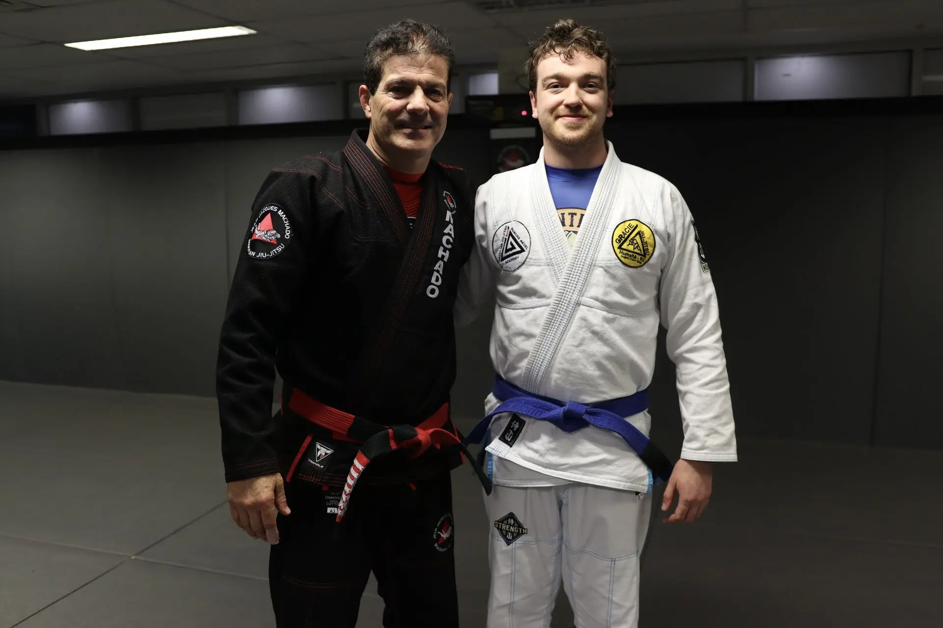 Jean-Jacques Machado, in a black gi with a red belt, stands next to a blue belt jiu-jitsu practitioner wearing a white gi with patches at Alpha Jiu-Jitsu Academy. Both are smiling and standing on the mats, emphasizing their camaraderie and dedication to Brazilian Jiu-Jitsu training.