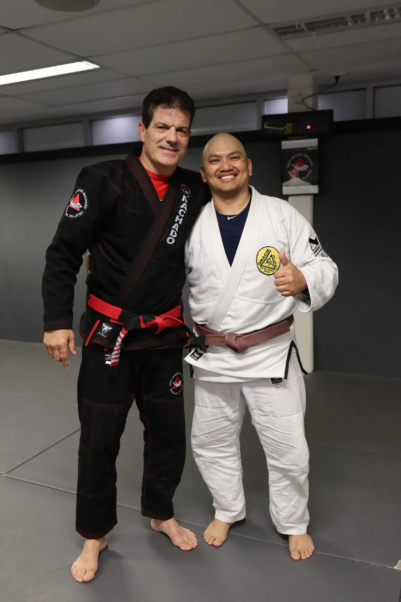 Jean-Jacques Machado, wearing a black gi and red belt, stands beside a smiling student in a white gi and brown belt at Alpha Jiu-Jitsu Academy. The student gives a thumbs-up gesture, expressing enthusiasm and camaraderie. The two are positioned in front of the academy’s training area, reflecting a sense of mentorship, respect, and achievement in Brazilian Jiu-Jitsu training.