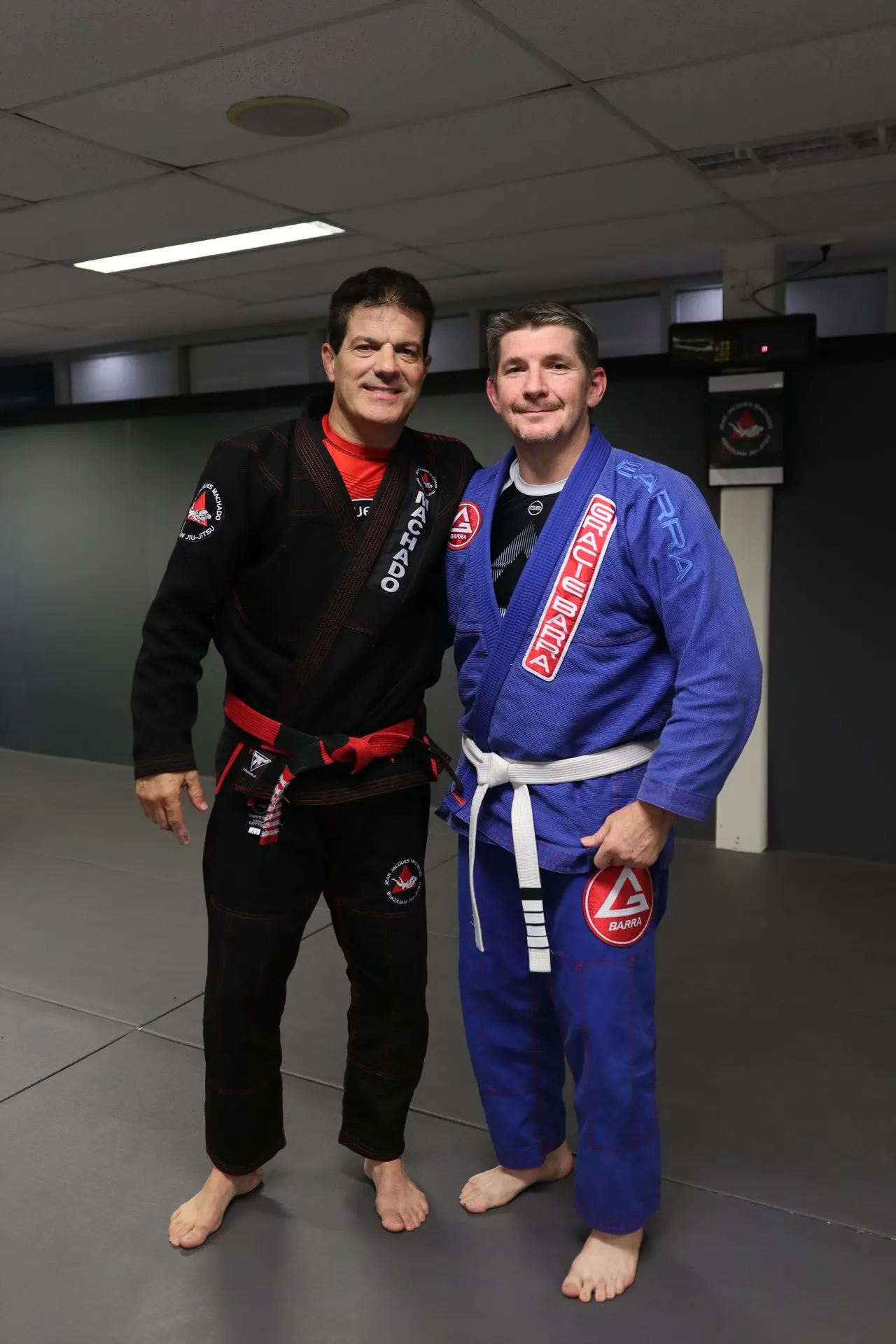 Jean-Jacques Machado standing with a white belt student wearing a blue Gracie Barra gi at Alpha Jiu Jitsu Academy, smiling together on the academy mat.