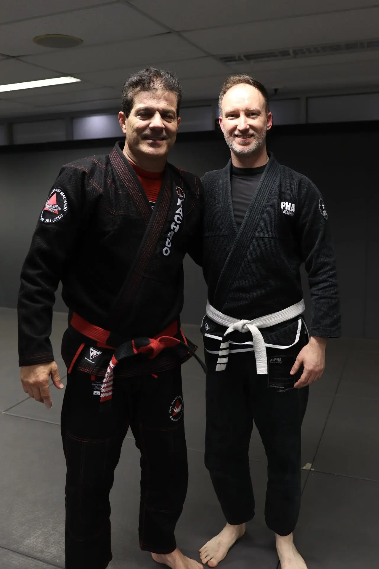 Jean-Jacques Machado, wearing a black gi and a red belt, poses alongside a white belt student in a black gi at Alpha Jiu-Jitsu Academy. Both are smiling and standing on the training mats, symbolizing camaraderie and dedication to Brazilian Jiu-Jitsu practice.