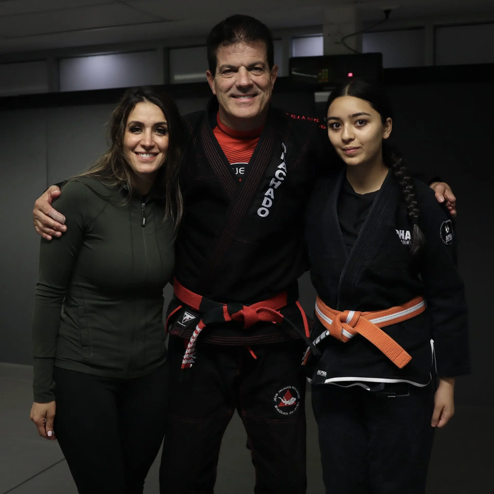 Jean-Jacques Machado, a legendary Brazilian Jiu-Jitsu figure, stands in the center with his arms around Marie Sales, Program Director, on the left, and a young female jiu-jitsu student with an orange belt on the right at Alpha Jiu Jitsu Academy.