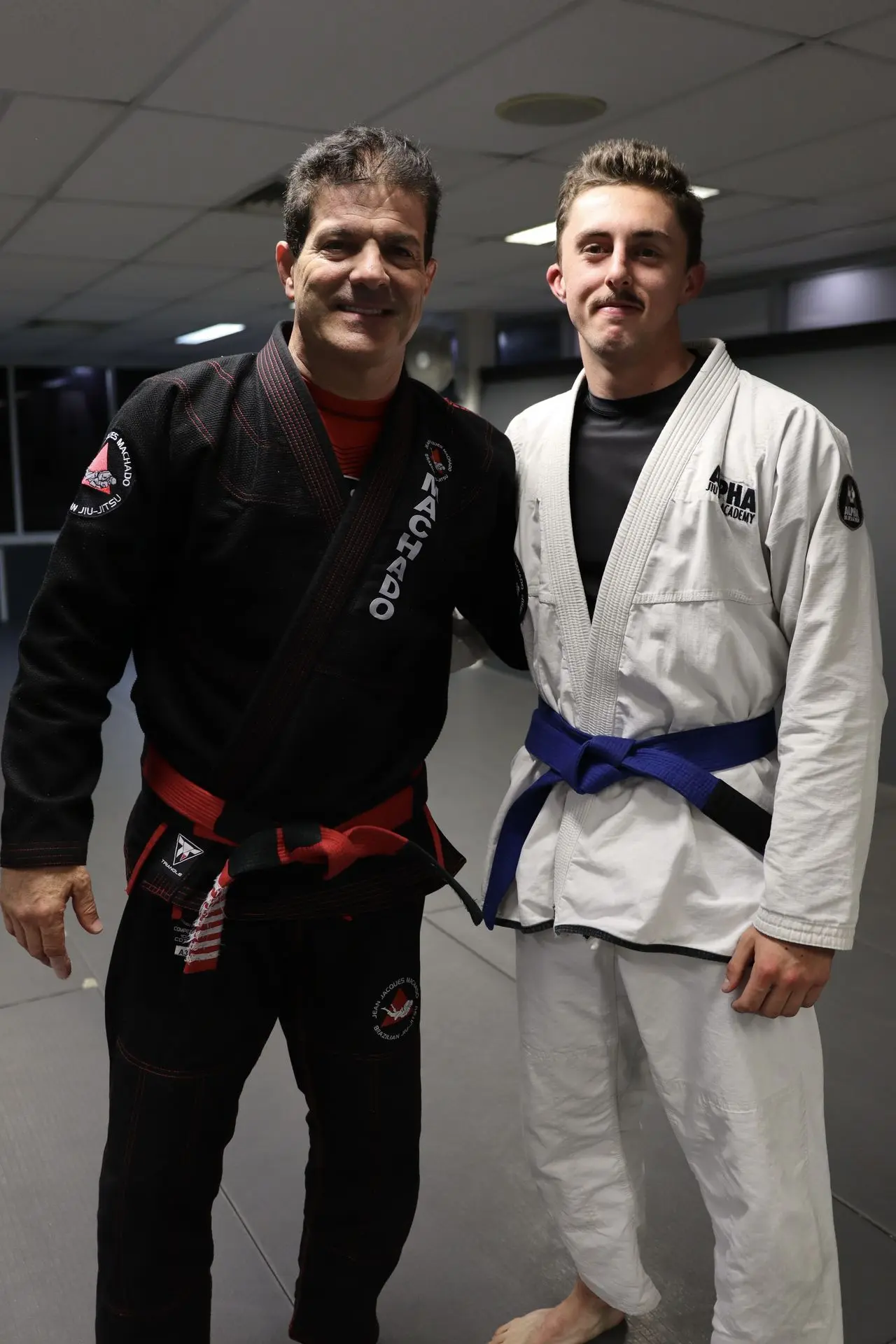 Jean-Jacques Machado, wearing a black gi and red belt, stands next to a blue belt Brazilian Jiu-Jitsu student dressed in a white gi. They are smiling for the photo, highlighting a moment of camaraderie and mentorship on the mats.