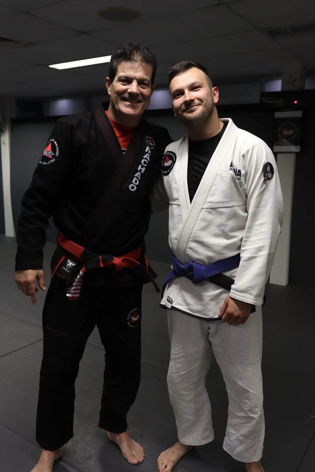 Jean-Jacques Machado, wearing a black gi with a red belt, stands with a smiling blue belt student in a white gi at Alpha Jiu-Jitsu Academy. The image captures the camaraderie and respect shared between instructor and student in the Brazilian Jiu-Jitsu community.