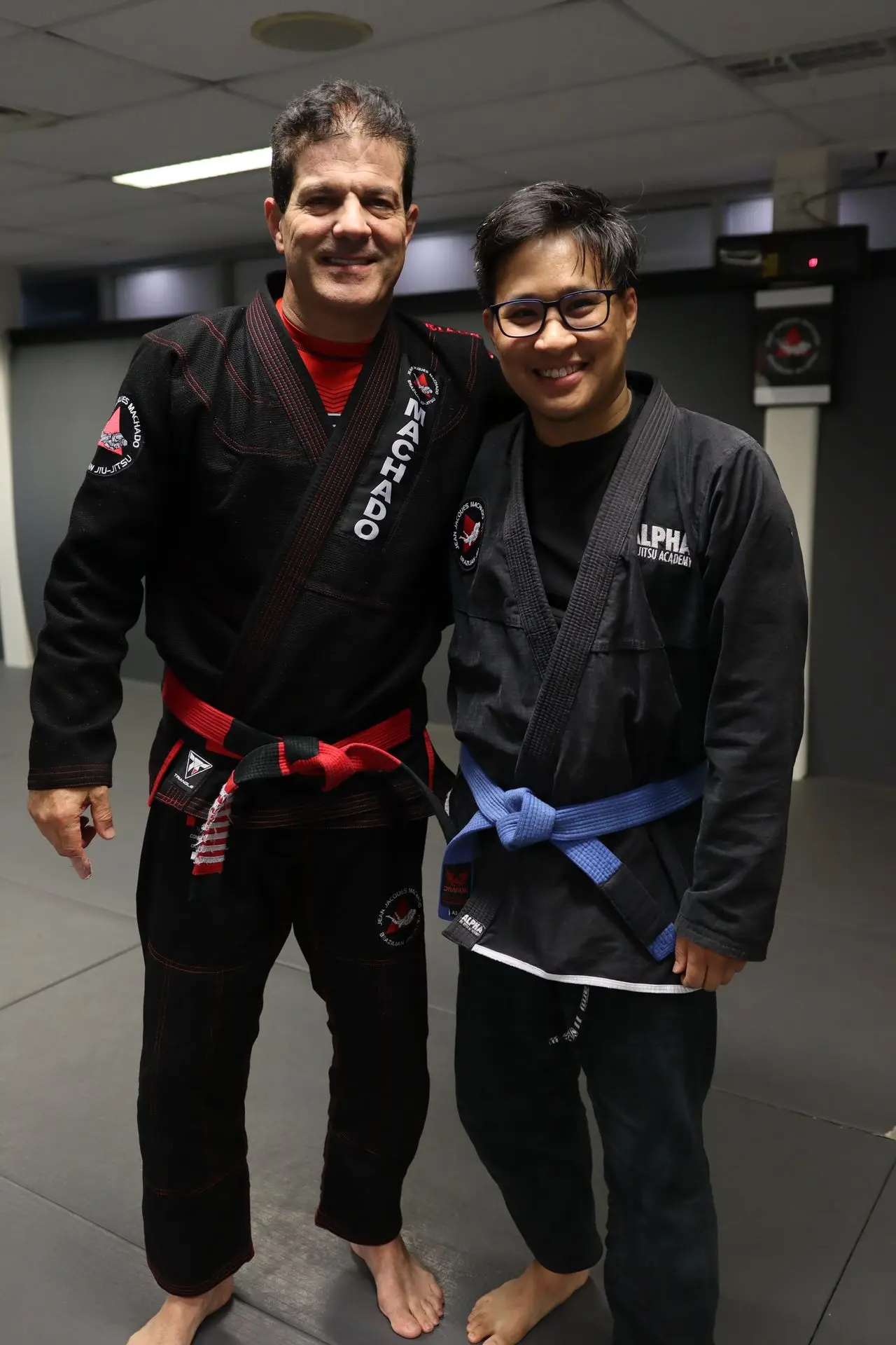 Jean-Jacques Machado with a blue belt student from Alpha Jiu Jitsu Academy, standing together in Brazilian Jiu-Jitsu gis on the academy mat.