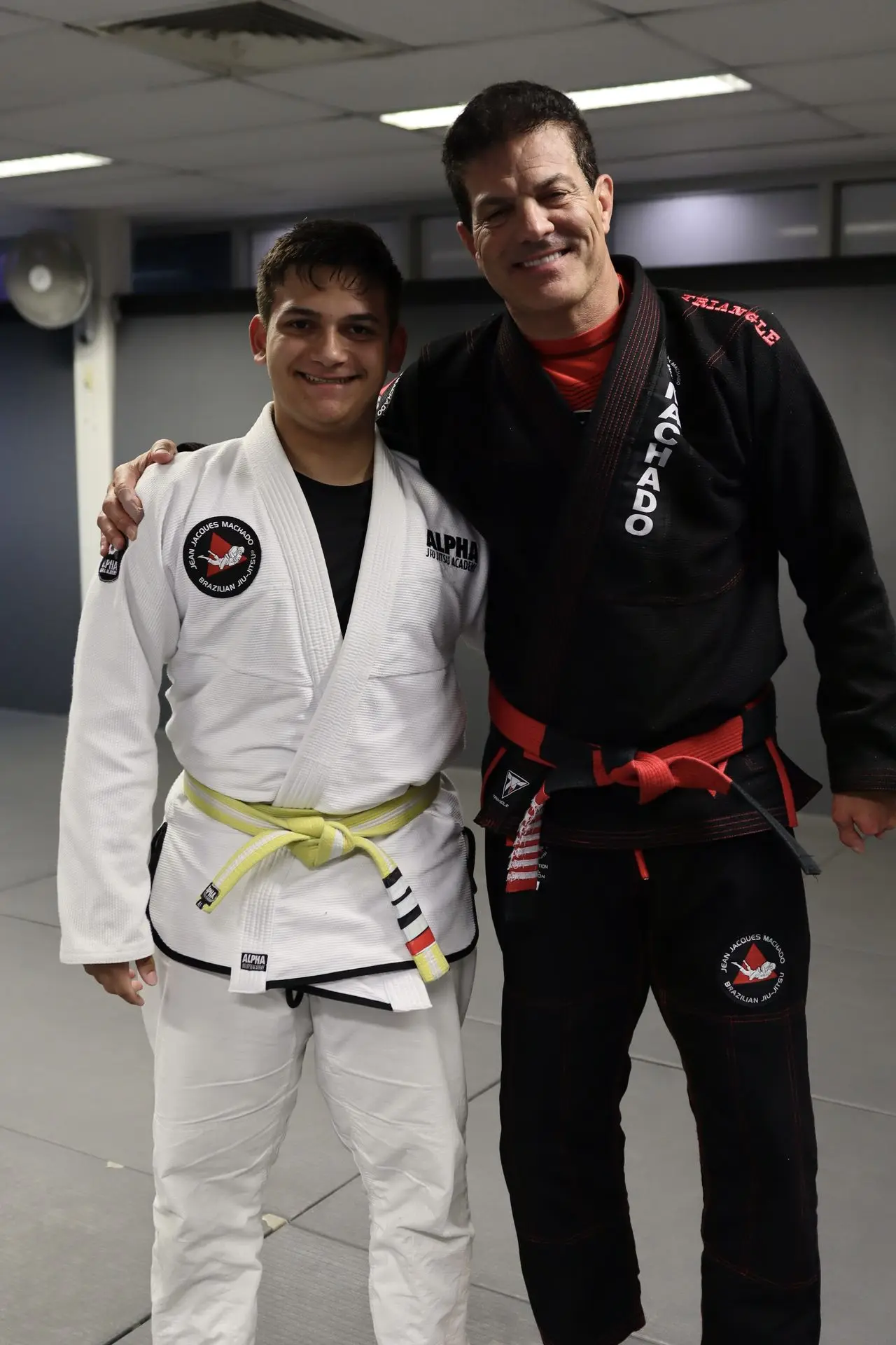 Jean-Jacques Machado posing with a BJJ student in a white gi with a yellow belt, both smiling at the Brazilian Jiu-Jitsu academy training facility.