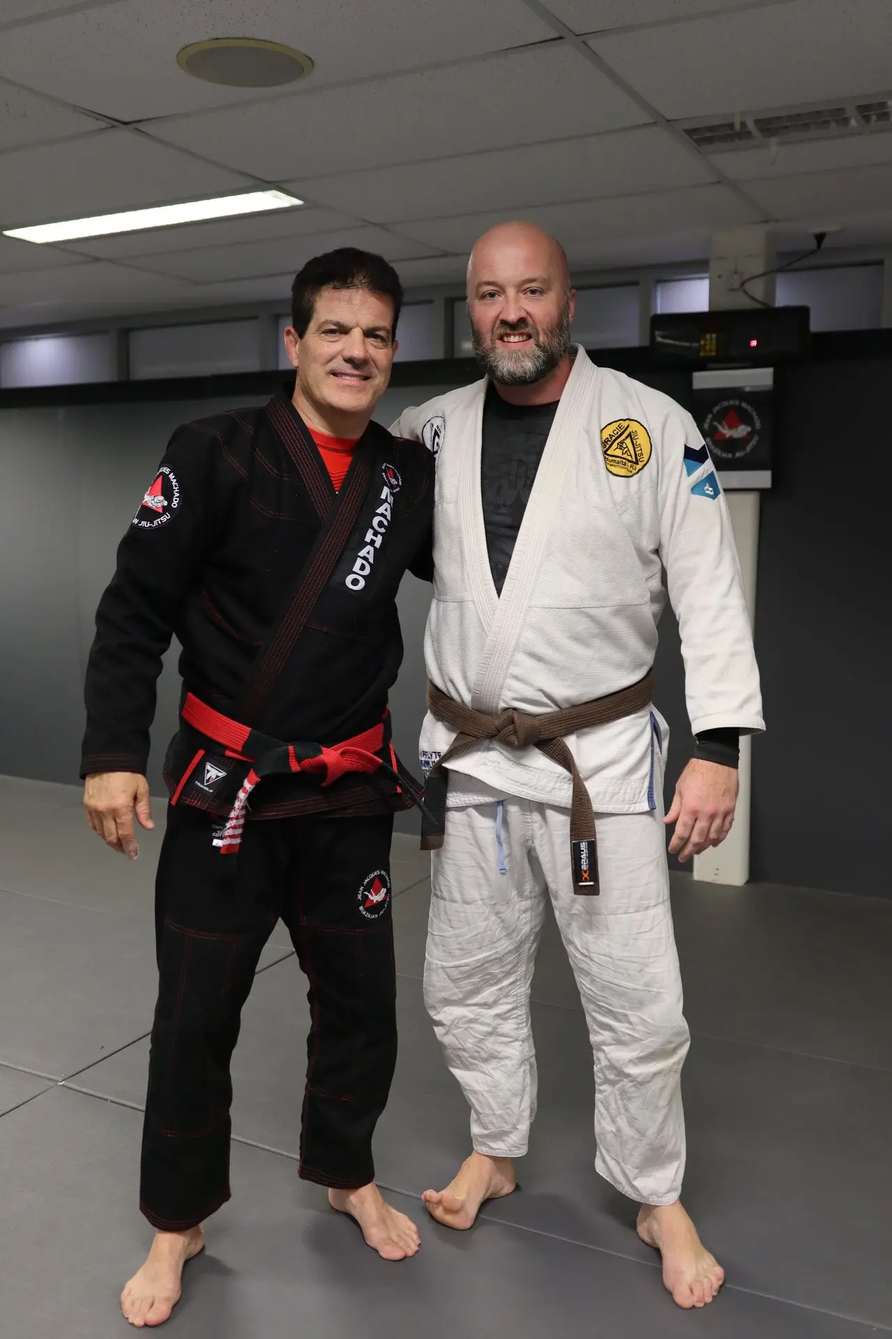 Jean-Jacques Machado, in a black gi and red belt, standing next to a student in a white gi and brown belt, both smiling at Alpha Jiu-Jitsu Academy. The student has a trimmed beard and stands proudly with Machado in front of the academy’s training area, reflecting mentorship, skill, and the spirit of camaraderie in Brazilian Jiu-Jitsu training.