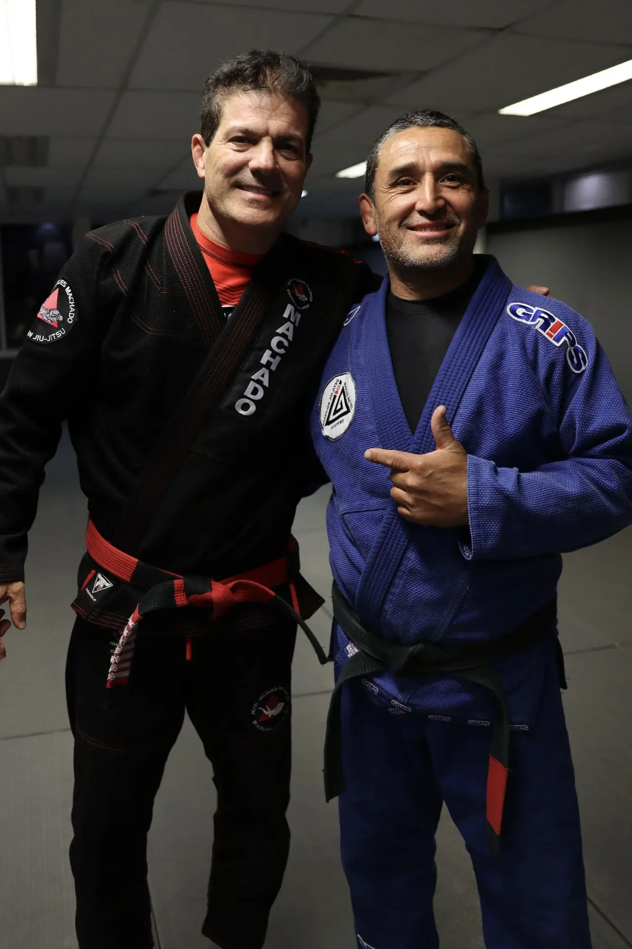 Jean-Jacques Machado, wearing a black gi with a red belt, stands next to a black belt BJJ practitioner in a blue gi. They are smiling and posing for a photo on the mats, capturing a memorable moment at a Brazilian Jiu-Jitsu event.