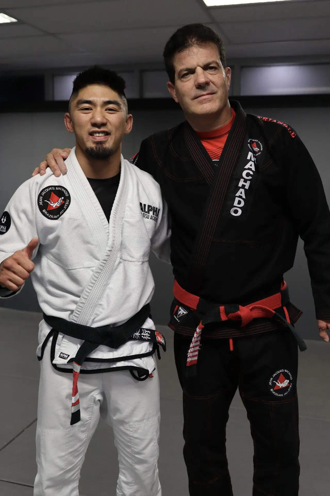 Professor Xavier Sales standing alongside Jean-Jacques Machado at Alpha Jiu Jitsu Academy, both wearing Brazilian Jiu-Jitsu gis, smiling after a training session.