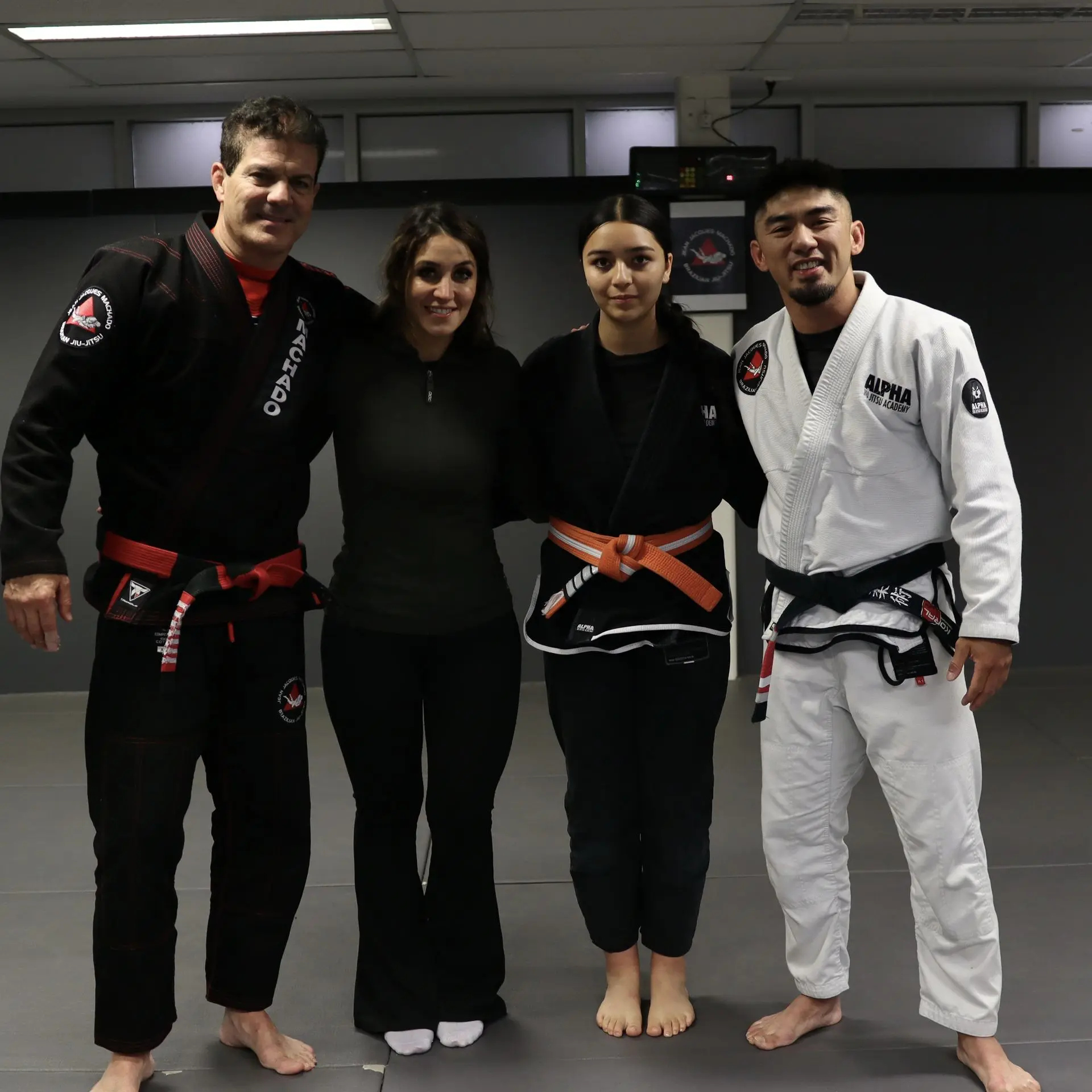 Group photo featuring Jean-Jacques Machado in a black gi with a red belt, standing next to Marie Sales, the Program Director, an orange belt student wearing a black gi, and Professor Xavier Sales in a white gi with a black belt. They are all posing together at Alpha Jiu-Jitsu Academy, reflecting camaraderie, discipline, and mentorship within Brazilian Jiu-Jitsu training.