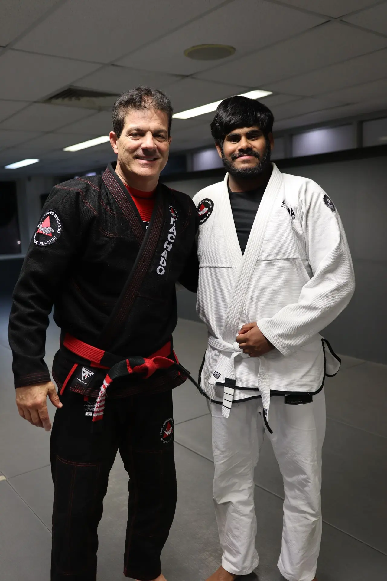 Jean-Jacques Machado, in a black gi with red belt, standing beside a white belt student in a white gi at Alpha Jiu Jitsu Academy, both smiling on the academy mats.