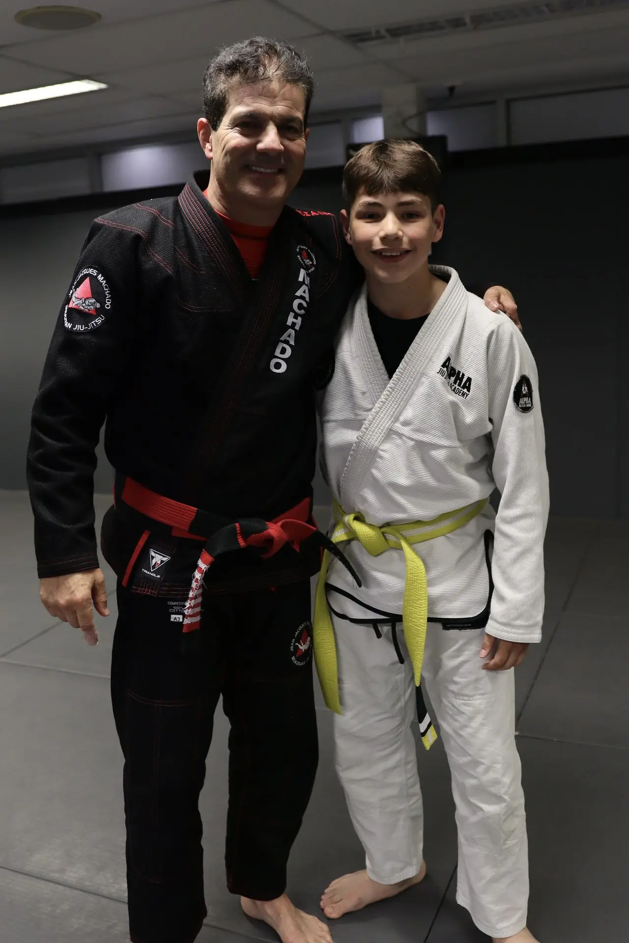 Jean-Jacques Machado, wearing a black gi with a red belt, stands next to a young student in a white gi with a yellow belt at Alpha Jiu-Jitsu Academy. They are smiling and posing together, representing the spirit of mentorship and growth in Brazilian Jiu-Jitsu training.