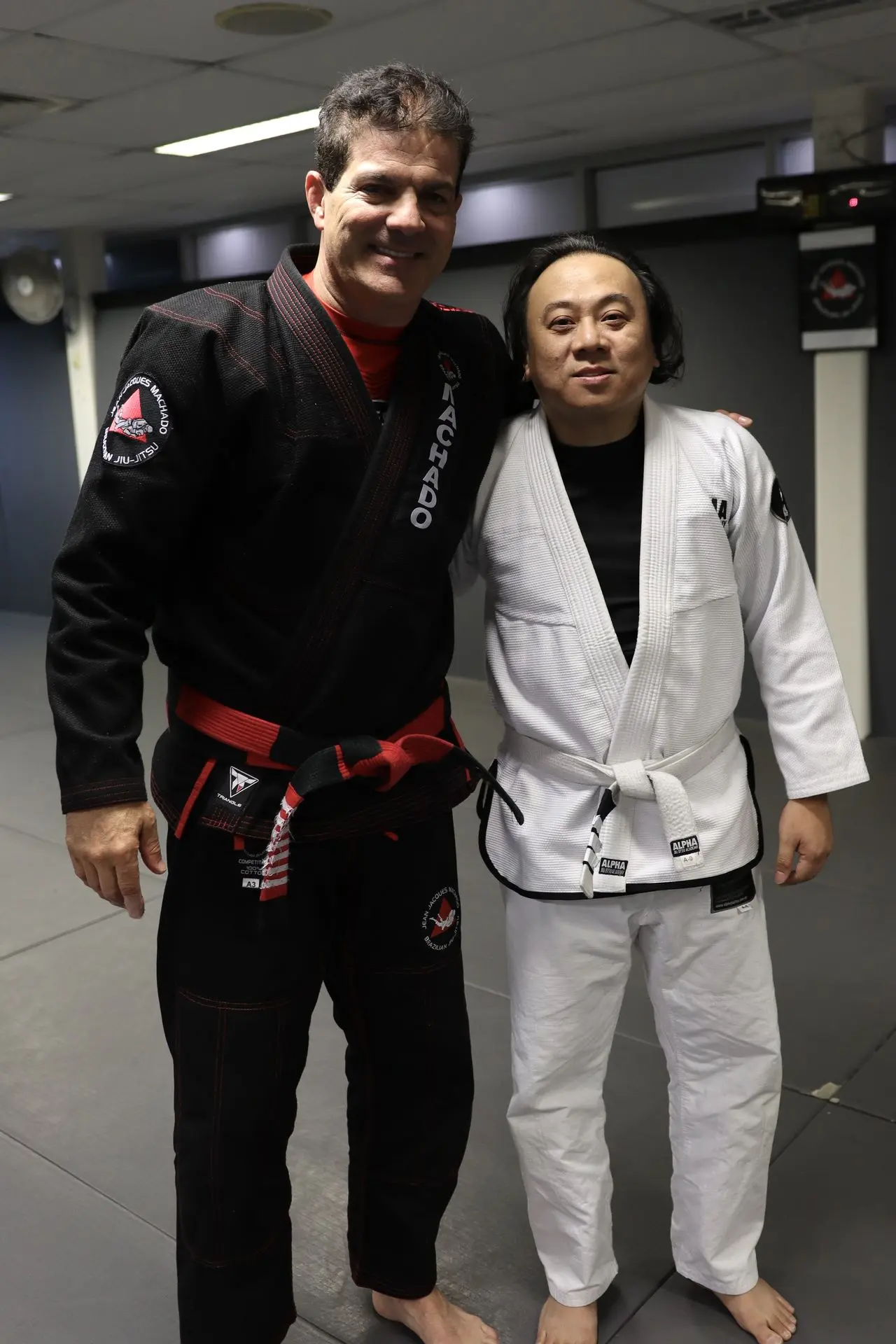 Jean-Jacques Machado posing with a Brazilian Jiu-Jitsu student wearing a white gi at the Alpha Jiu Jitsu Academy training facility. Both are smiling, standing side by side.