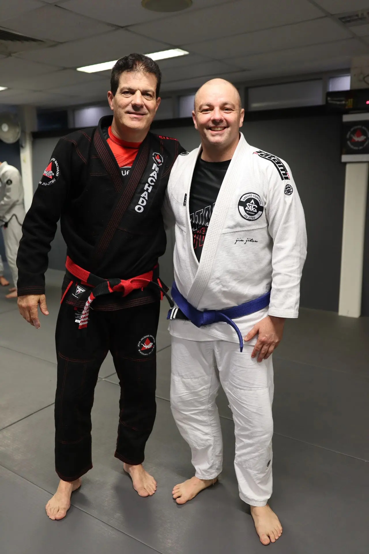 Jean-Jacques Machado, wearing a black gi and red belt, stands next to a BJJ practitioner in a white gi with a blue belt at Alpha Jiu Jitsu Academy. Both are smiling, standing on the training mats during a martial arts event.