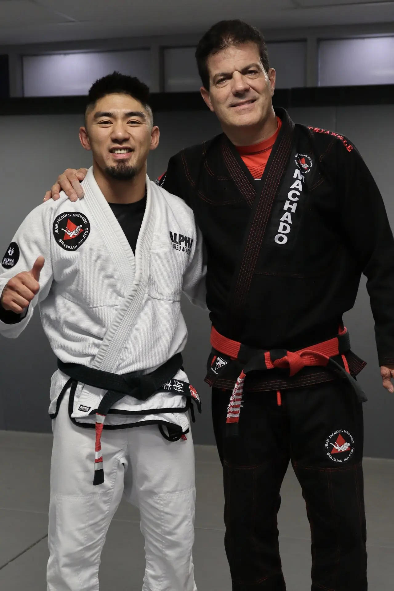 Professor Xavier Sales, a Brazilian Jiu-Jitsu instructor from Alpha Jiu Jitsu Academy, standing next to BJJ legend Jean-Jacques Machado. Both are wearing their gi, smiling for the camera during a martial arts event.