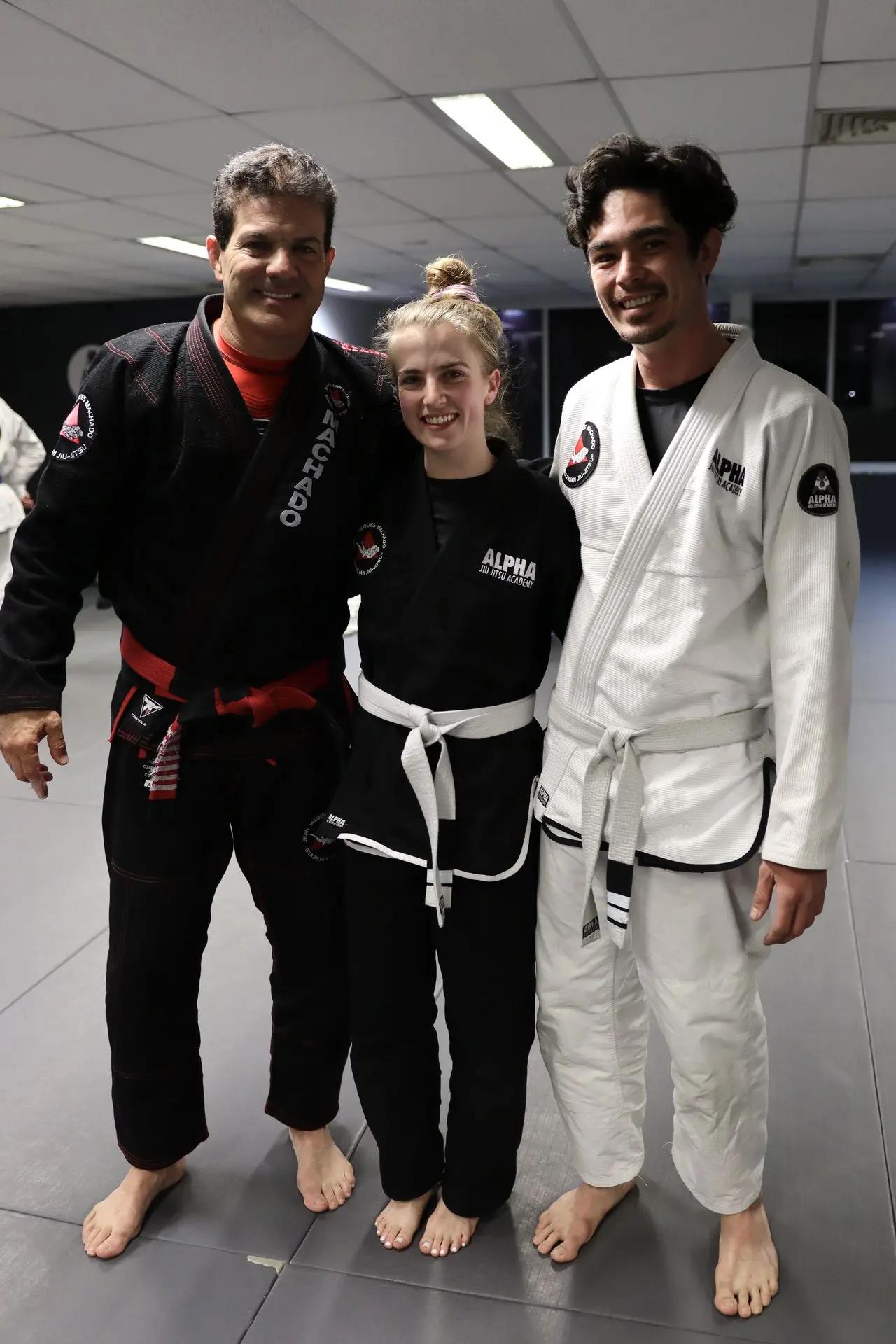 Jean-Jacques Machado, wearing a black gi with a red belt, stands alongside two white belt Brazilian Jiu-Jitsu students, one in a black gi and the other in a white gi, at Alpha Jiu-Jitsu Academy. They are smiling and posing together on the training mats, showcasing the sense of community and dedication in the martial arts journey.