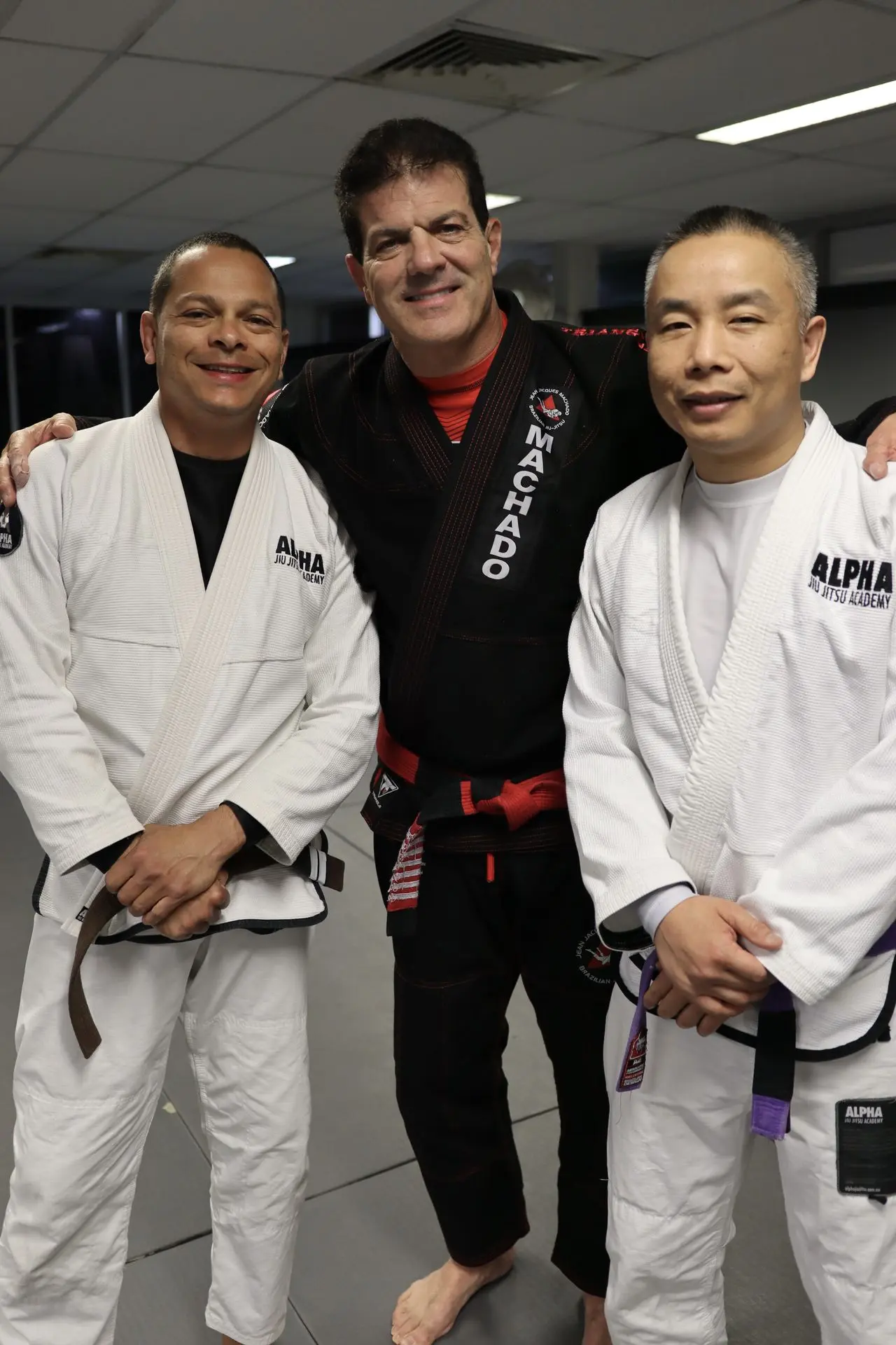 Jean-Jacques Machado standing and smiling with two students from Alpha Jiu Jitsu Academy, all in Brazilian Jiu-Jitsu gis, posing together during a training session.