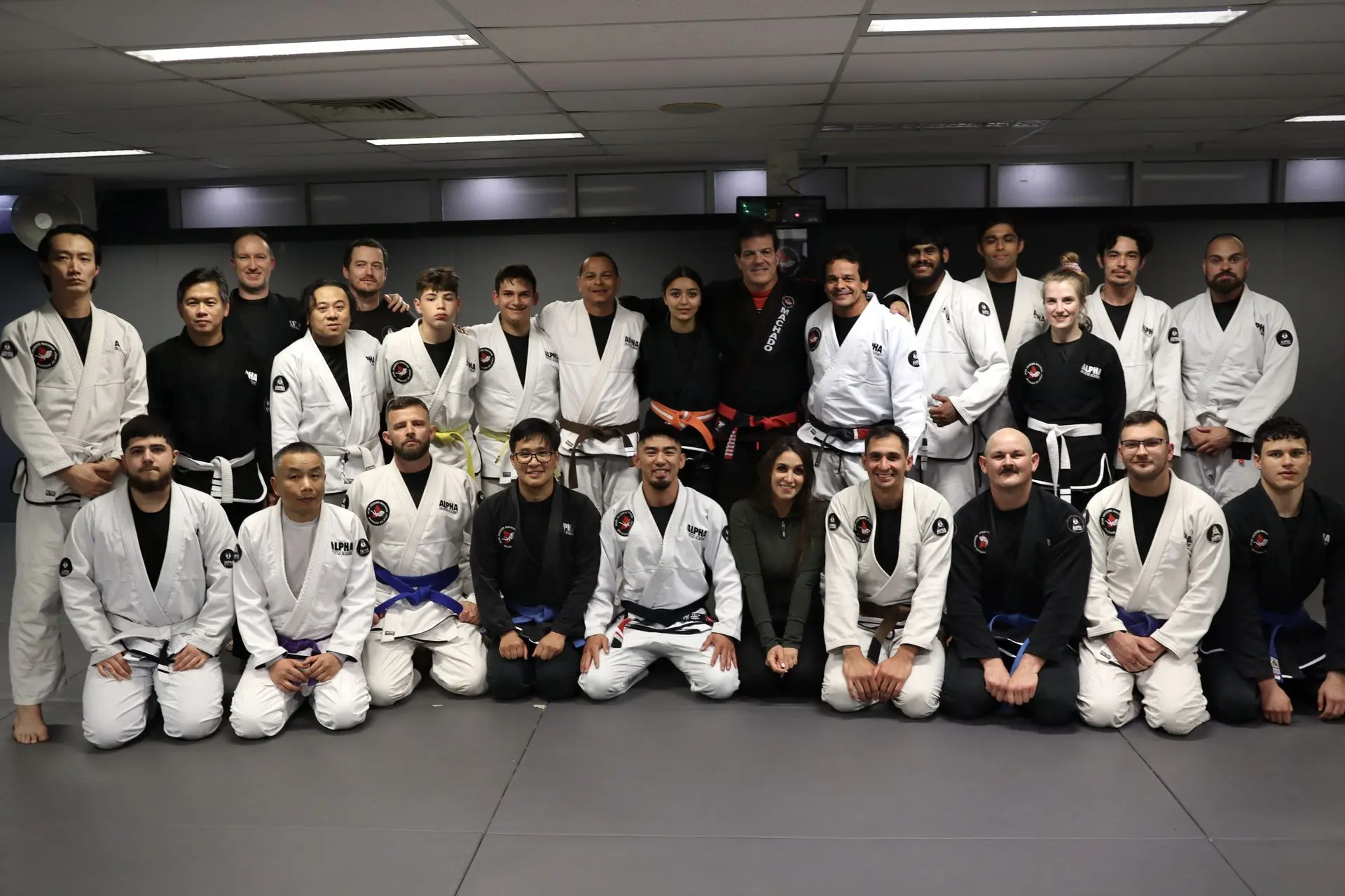 A large group of students and instructors at Alpha Jiu-Jitsu Academy pose for a group photo alongside Jean-Jacques Machado, who stands at the center wearing a black gi and red belt. The participants, dressed in a mix of white and black gis, display various belt ranks, reflecting their camaraderie and commitment to Brazilian Jiu-Jitsu training. The group showcases unity, diversity, and dedication within the academy's community.