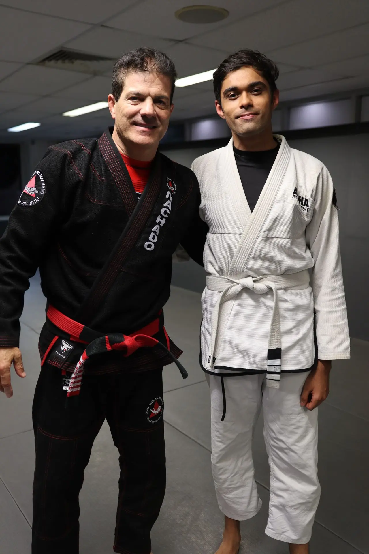 Jean-Jacques Machado, wearing a black gi and red belt, poses with a white belt student at Alpha Jiu-Jitsu Academy. The pair stand side by side on the training mat, symbolizing mentorship and growth within Brazilian Jiu-Jitsu. The student wears a white gi with the academy's branding and smiles confidently, reflecting the supportive and empowering atmosphere of the training session.