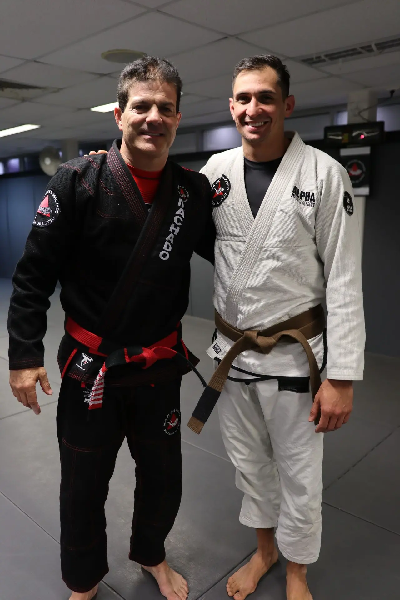 Jean-Jacques Machado poses with a brown belt practitioner from Alpha Jiu-Jitsu Academy. The pair stand on the training mat, demonstrating camaraderie and respect within the BJJ community. The brown belt student smiles proudly while wearing a white gi with Alpha Jiu-Jitsu Academy branding. The image reflects a moment of achievement and connection in Brazilian Jiu-Jitsu training.