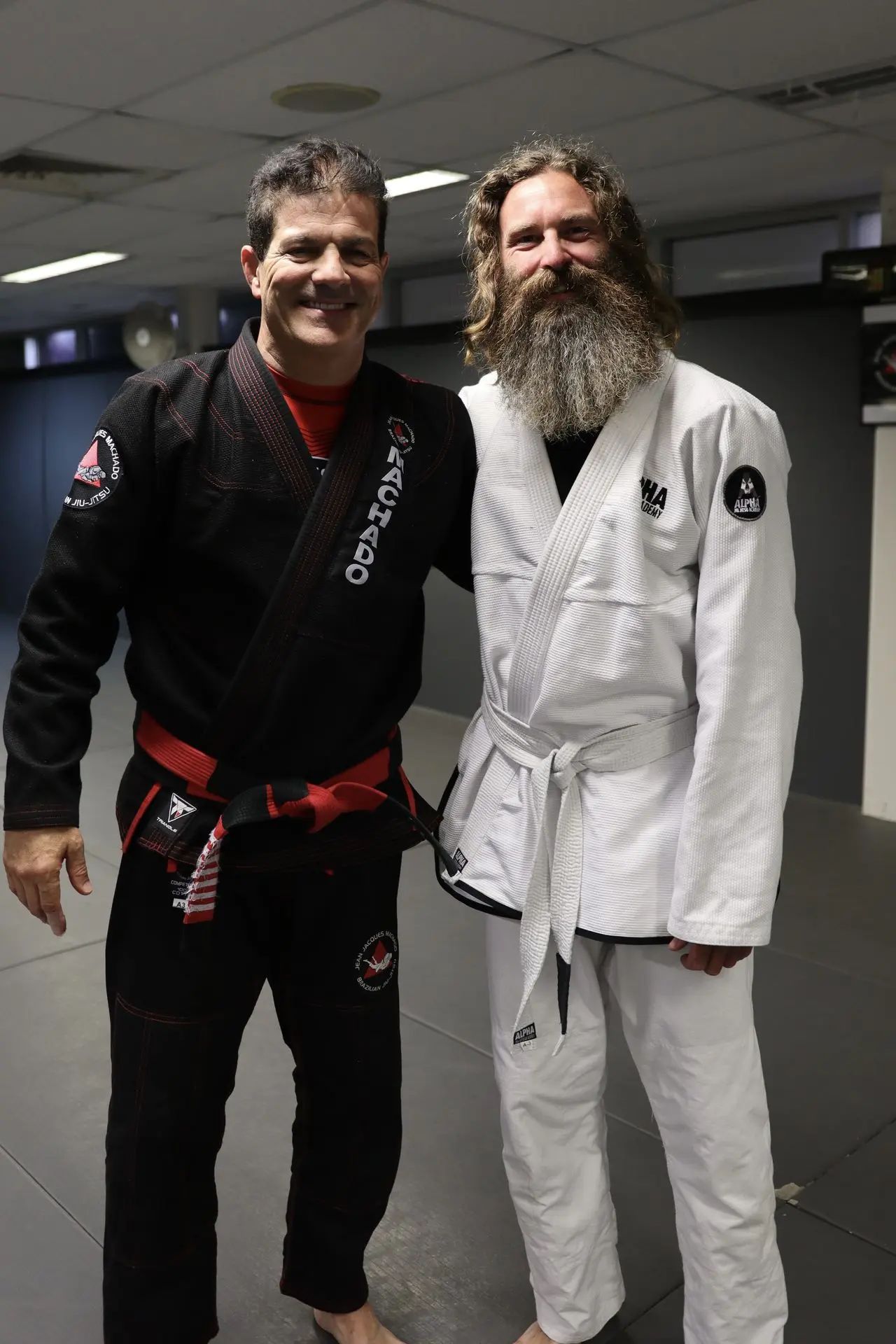 Jean-Jacques Machado, in a black gi and red belt, stands next to a white belt practitioner with a long beard, wearing a white gi at Alpha Jiu-Jitsu Academy. Both are smiling on the mat, highlighting their mutual respect and dedication to Brazilian Jiu-Jitsu training.