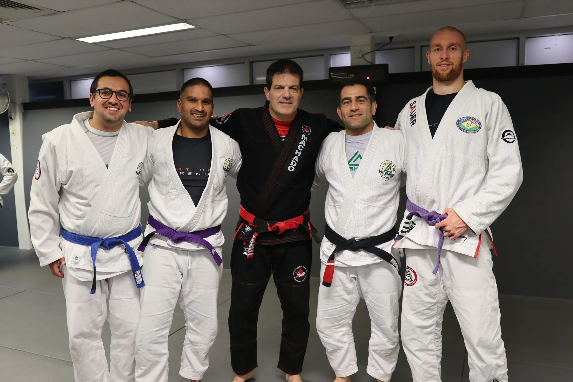 Jean-Jacques Machado stands in the center, flanked by four Brazilian Jiu-Jitsu practitioners wearing white gis with various belt colors, including purple and blue, at Alpha Jiu-Jitsu Academy. The group poses together, demonstrating camaraderie, discipline, and mastery within the martial arts setting.