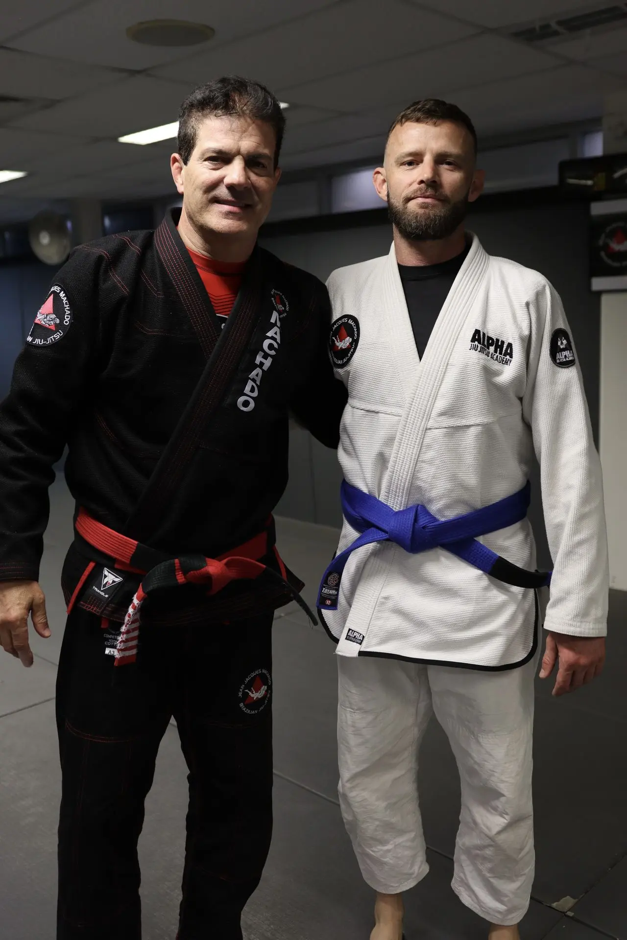 Jean-Jacques Machado, wearing a black gi with red belt, standing next to a blue belt BJJ practitioner dressed in a white gi at Alpha Jiu Jitsu Academy. Both are smiling and standing side by side.