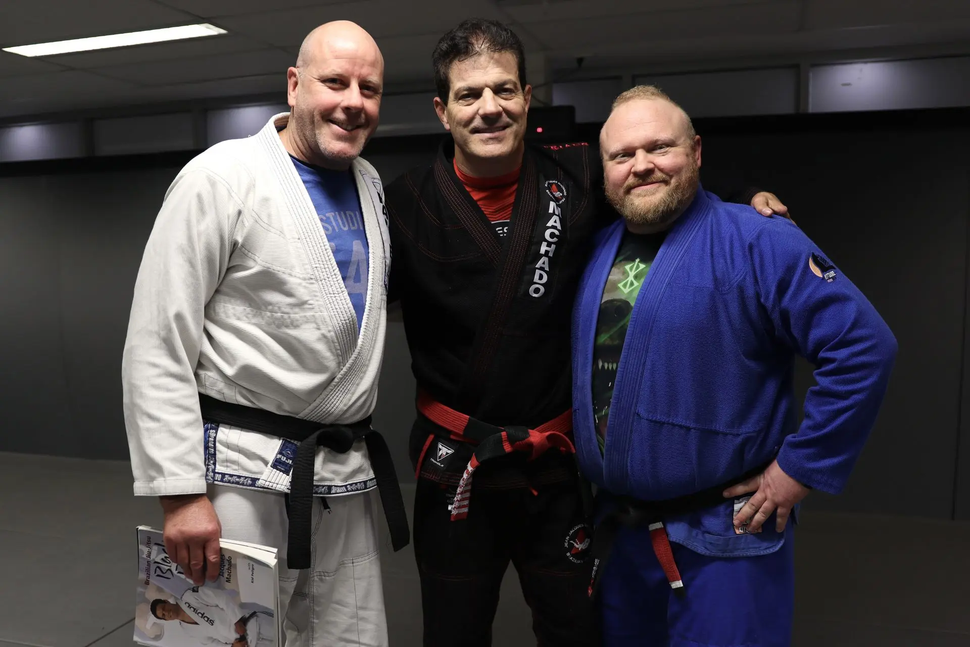 Jean-Jacques Machado, wearing a black gi with a red belt, standing between two BJJ practitioners. The practitioner on the left is wearing a white gi and holding a book, while the practitioner on the right is wearing a blue gi. All three are smiling and standing together at Alpha Jiu Jitsu Academy.