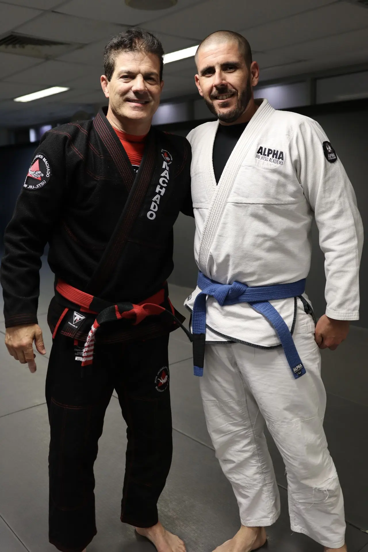 Jean-Jacques Machado, wearing a black gi with a red belt, standing next to a BJJ practitioner in a white gi with a blue belt. They are standing side by side at Alpha Jiu Jitsu Academy.