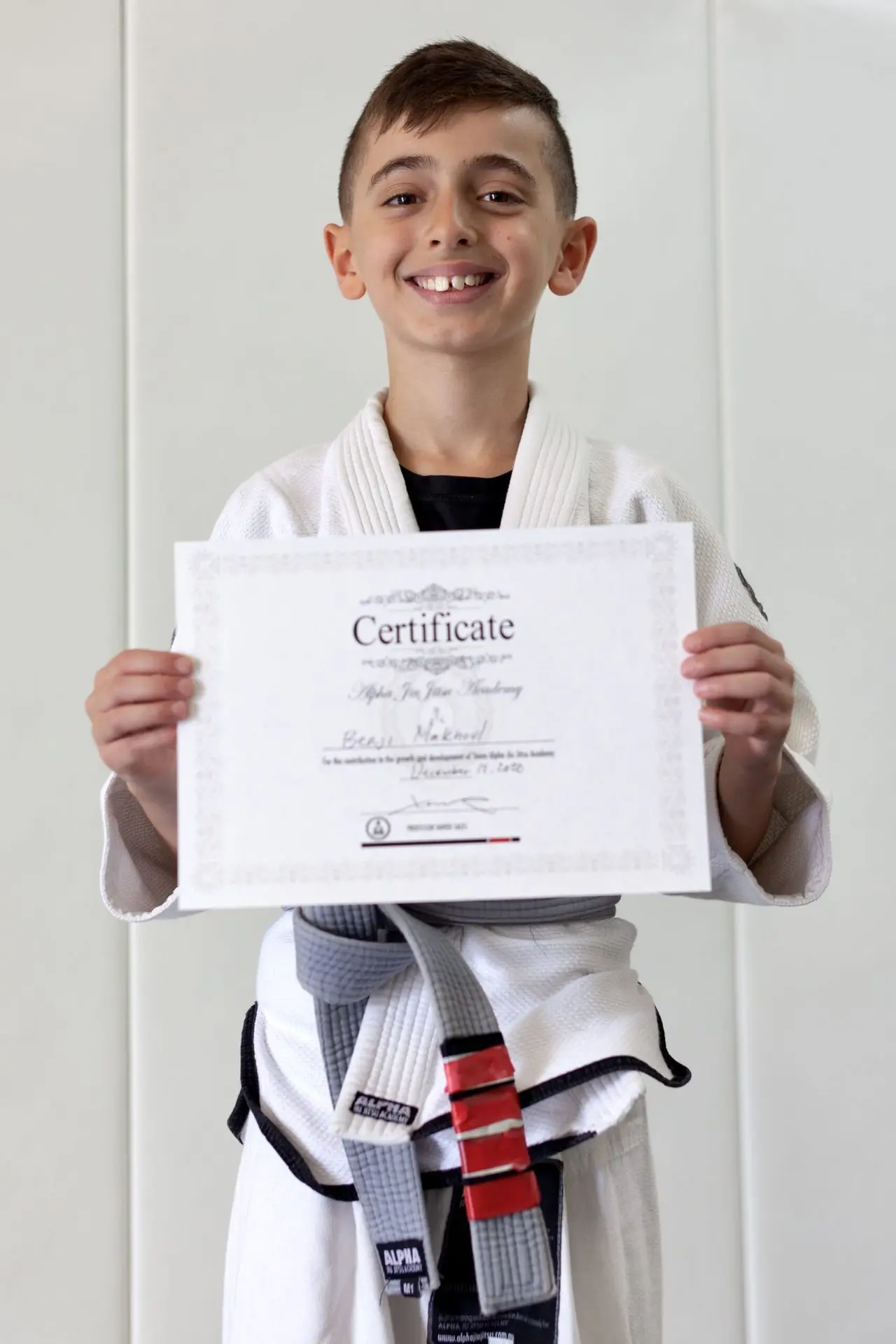 Beau Makoui, a young student at Alpha Jiu-Jitsu Academy, proudly holding his certificate of achievement while wearing a white gi and grey belt. Beau is smiling confidently, showcasing his accomplishment and dedication to training at the academy. The certificate bears his name, marking a milestone in his jiu-jitsu journey. The background features a plain wall, highlighting Beau's bright expression.