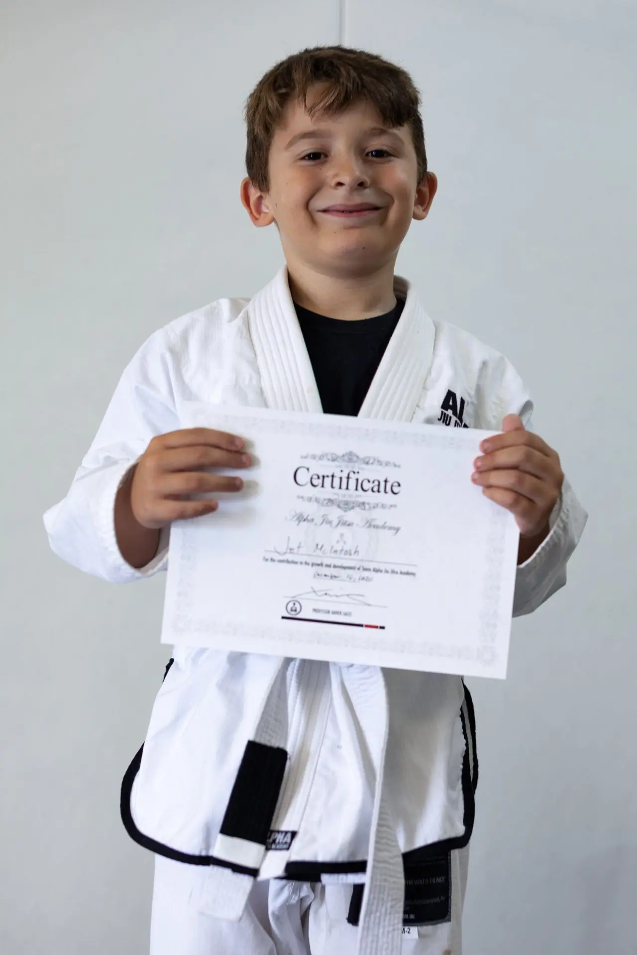 Jet McIntosh, a young student at Alpha Jiu-Jitsu Academy, proudly holding his graduation certificate. He is smiling, wearing a white gi with a black belt, and showing off his achievement. The certificate, featuring Jai’s name, celebrates his progress and dedication at the academy. The background is blurred to focus on Jai and his accomplishment.