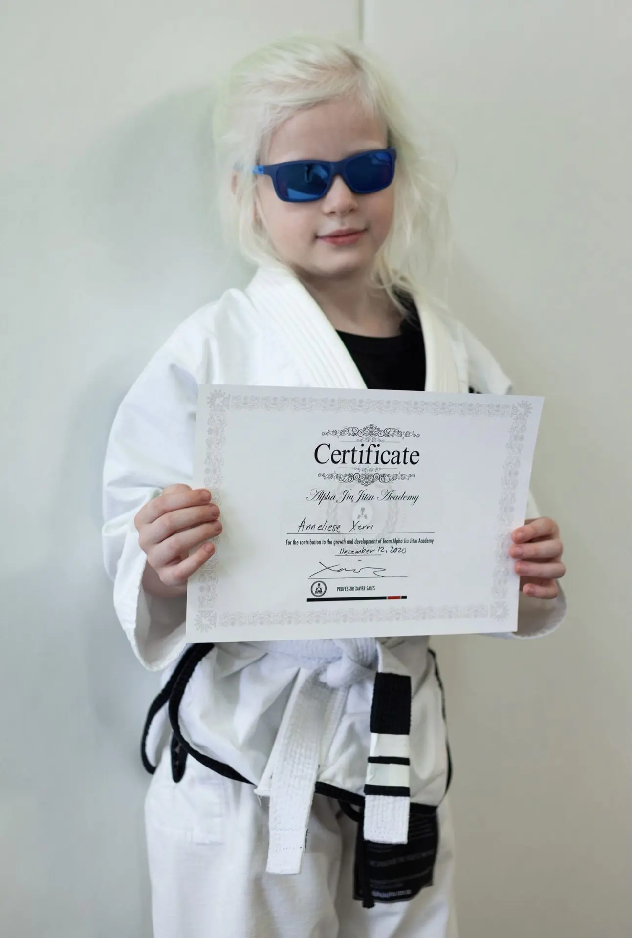 Annelese Xerri, a young student at Alpha Jiu-Jitsu Academy, wearing a white gi with a black belt, holds her graduation certificate. She is smiling and wearing blue sunglasses, standing proudly as she displays her achievement. The certificate, featuring her name, recognizes her progress and dedication within the academy. The background is simple and allows Annelese and her certificate to be the focal points of the image.