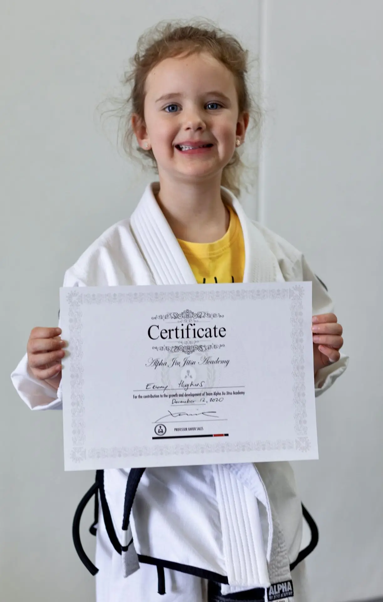 Ebony Hughes, a young student at Alpha Jiu-Jitsu Academy, proudly holds her graduation certificate while wearing a white gi over a yellow shirt. She smiles brightly, celebrating her accomplishment. The certificate, bearing her name, marks her progress and dedication within the program. The simple background emphasizes Ebony and her achievement.