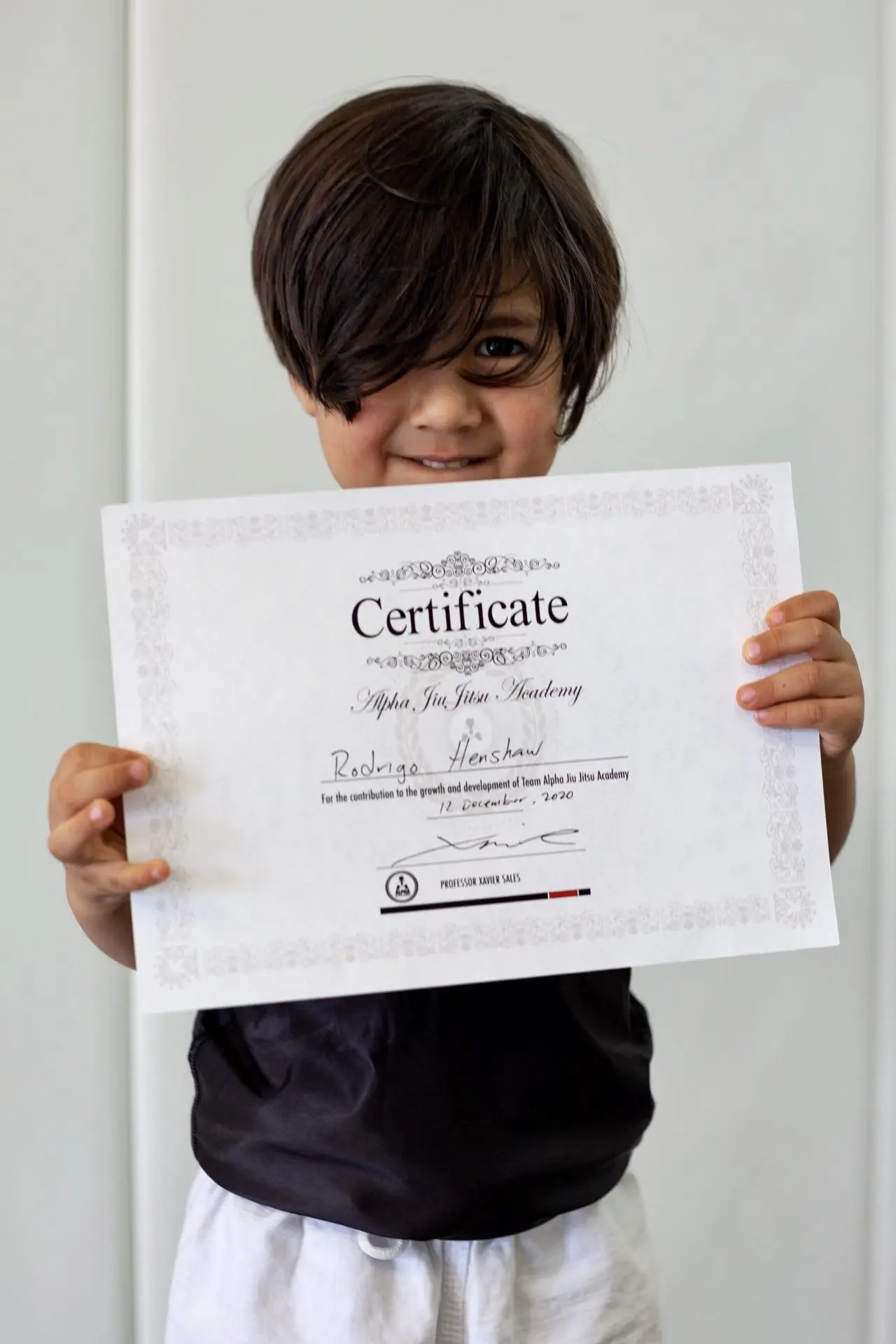 Rodrigo Henshaw, a young student at Alpha Jiu-Jitsu Academy, smiles while holding his certificate of achievement. He wears a black shirt and white gi pants, proudly showcasing his milestone. The certificate displays his name, marking his accomplishments and growth at the academy. The image captures Rodrigo's joyful expression, highlighting his dedication and progress.